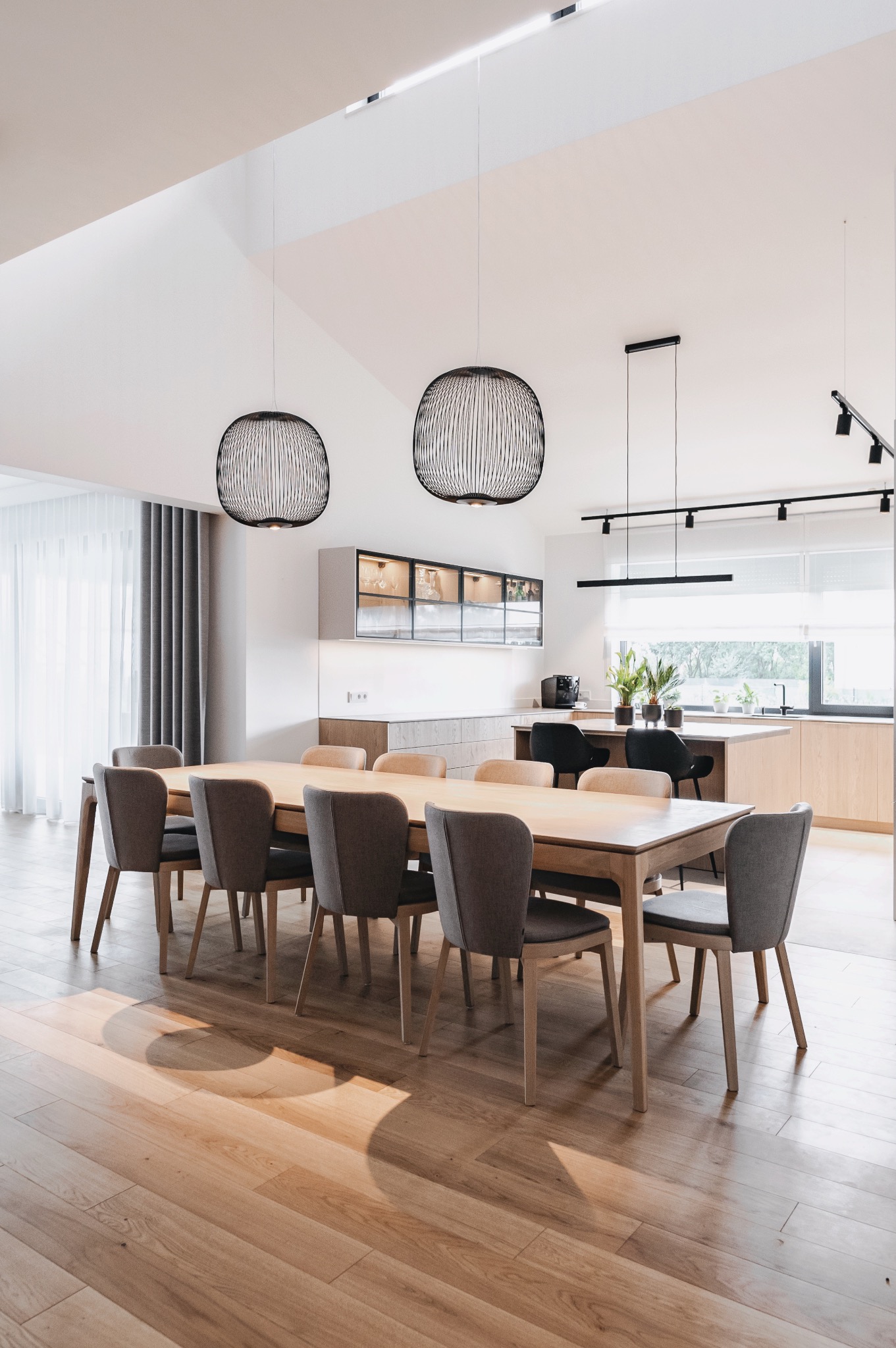 Jasna jadalnia z drewnianym stołem, szarymi krzesłami i nowoczesnymi lampami wiszącymi. W tle widoczna kuchnia z drewnianymi frontami i przeszklonymi szafkami.