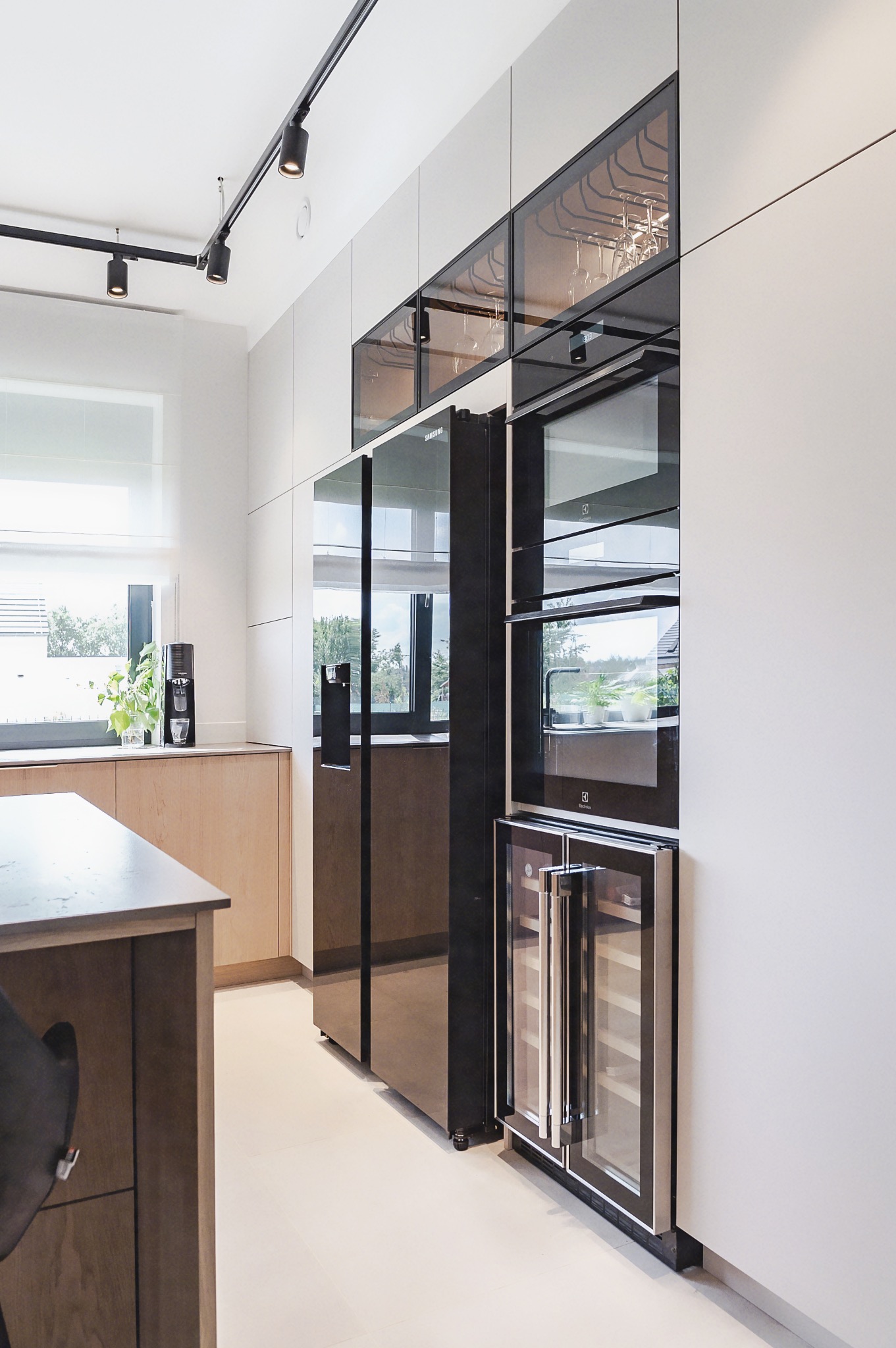 Nowoczesna kuchnia z zabudową w odcieniach bieli i drewna, czarny sprzęt AGD, oświetlenie szynowe i wyspa kuchenna z blatem w kolorze grafitowym.