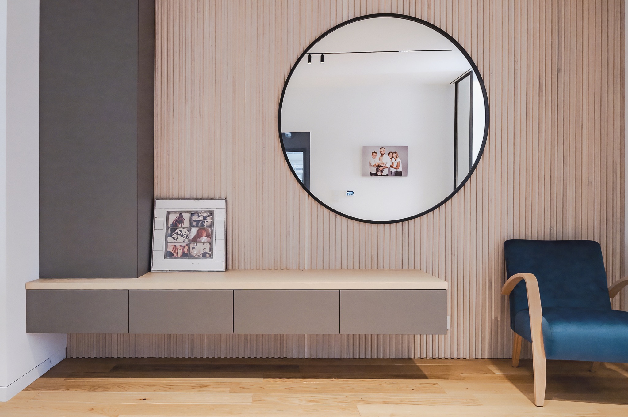 Nowoczesna aranżacja wnętrza: wisząca szafka z szufladami w kolorze taupe, okrągłe lustro i fotel na tle ściany z drewnianych paneli. Minimalizm i elegancja.
