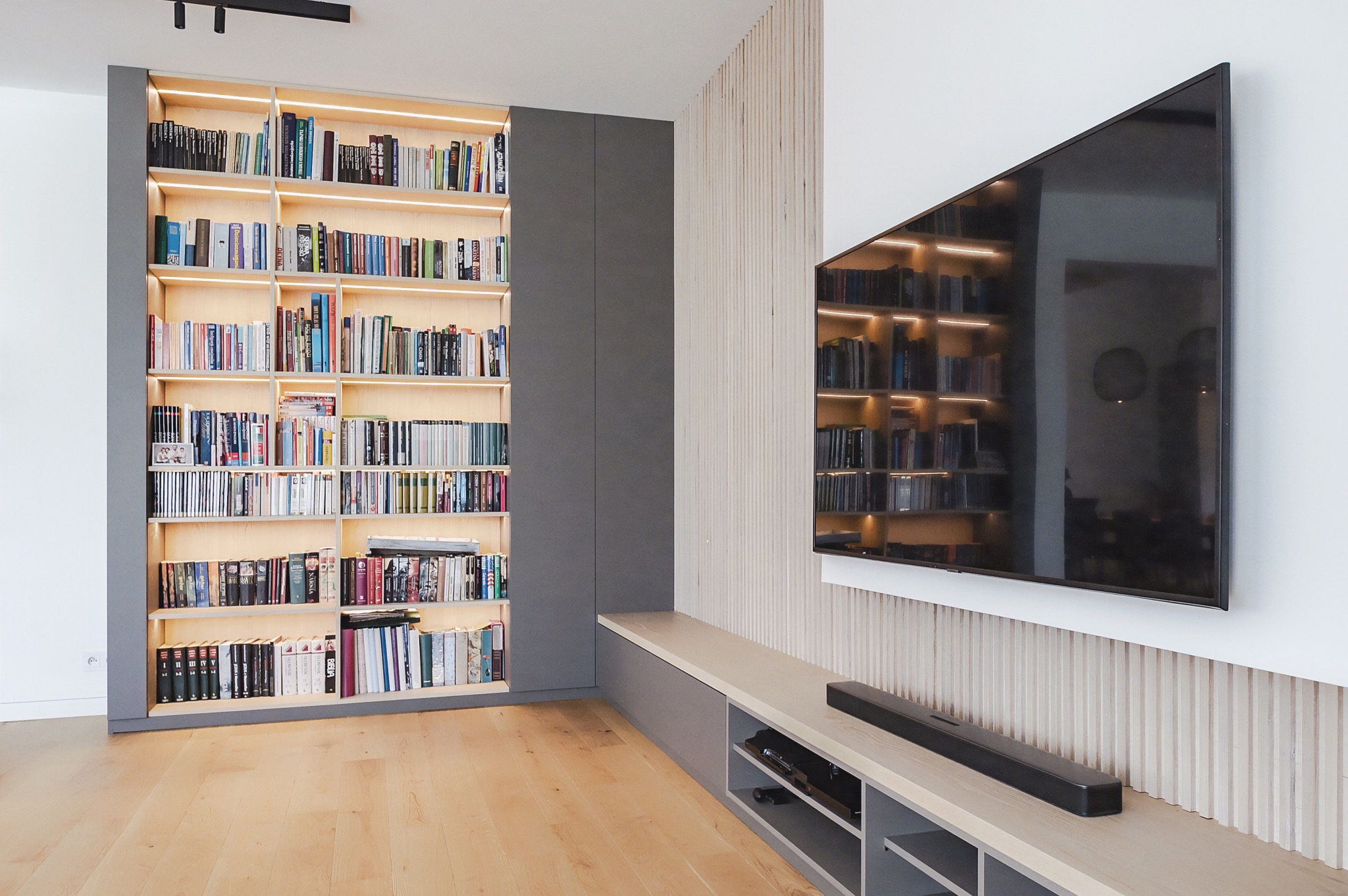 Nowoczesna szafa wnękowa z podświetlanymi półkami na książki, obok ściana z panelem dekoracyjnym i telewizorem. Jasna podłoga.