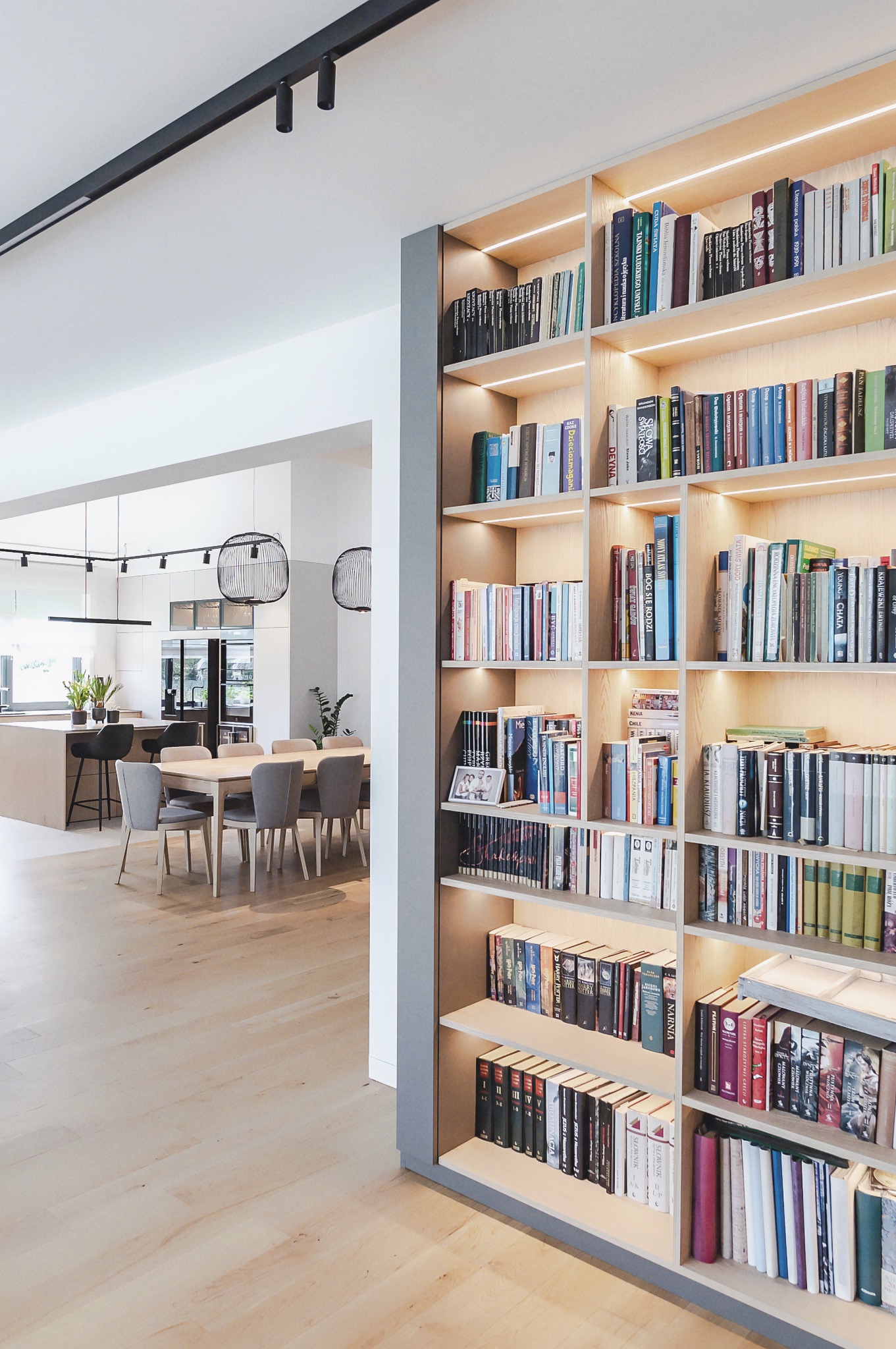 Regał na książki z oświetleniem LED, widoczny z perspektywy salonu z otwartą kuchnią. Jasne drewno i szare elementy wykończeniowe. Minimalistyczny design.