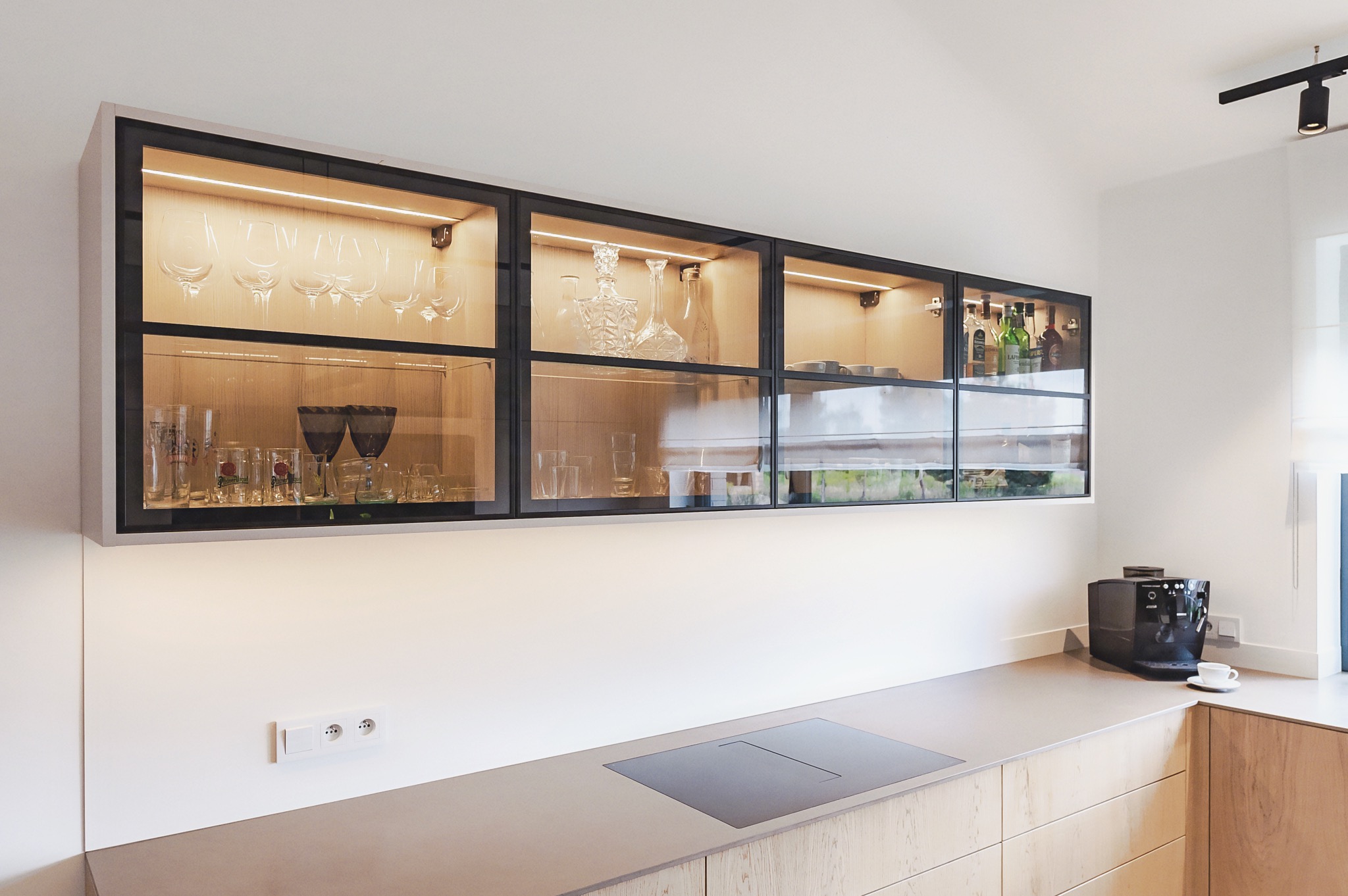 Nowoczesna kuchnia: przeszklone szafki wiszące z czarnymi ramami, podświetlane LED, eksponujące szkło i naczynia. Jasne drewno i blat, minimalistyczny design.