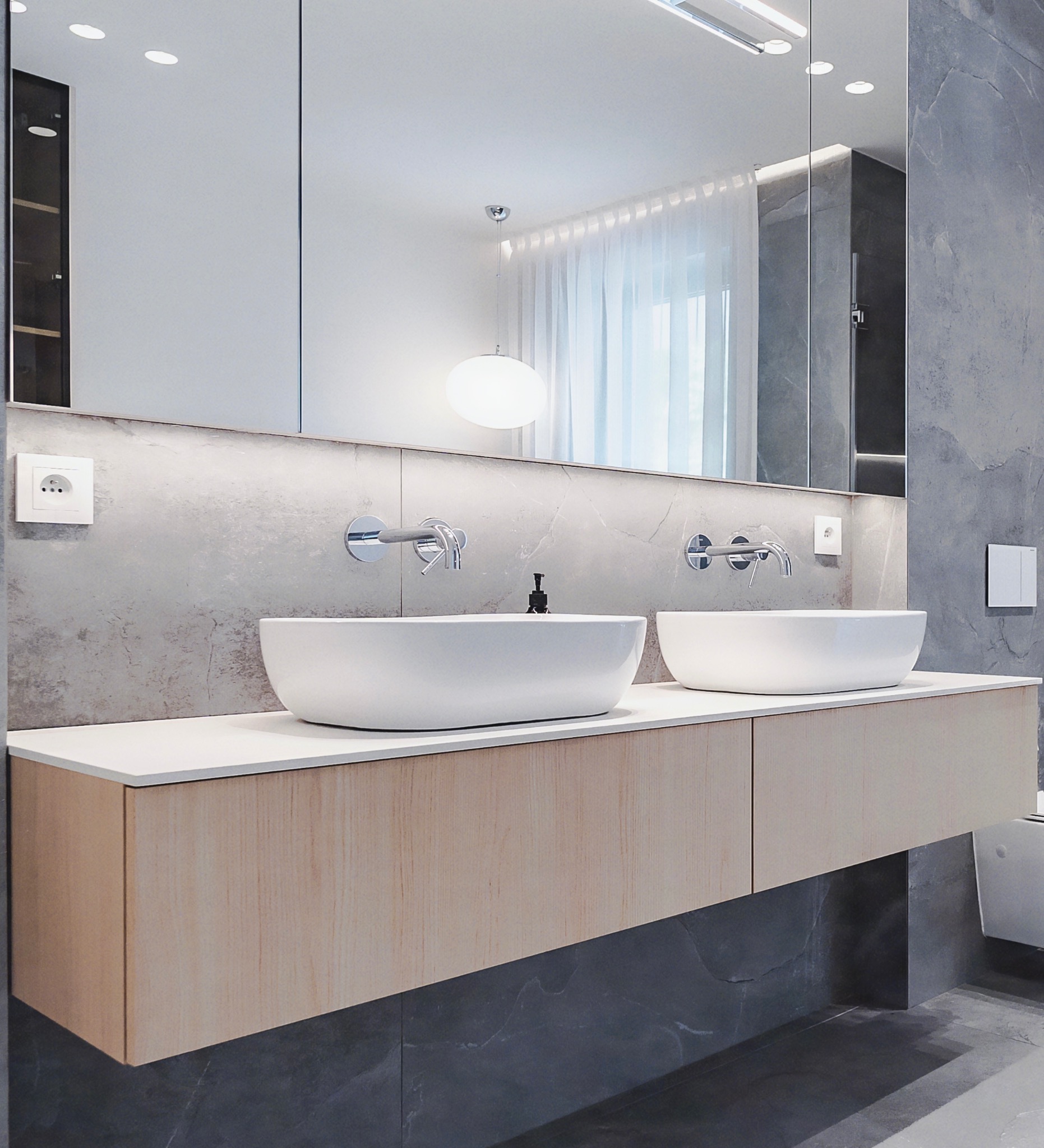 Nowoczesna łazienka z dwoma owalnymi umywalkami nablatowymi i szafką pod umywalkę z jasnego drewna na tle ściany z szarego kamienia i dużego lustra.