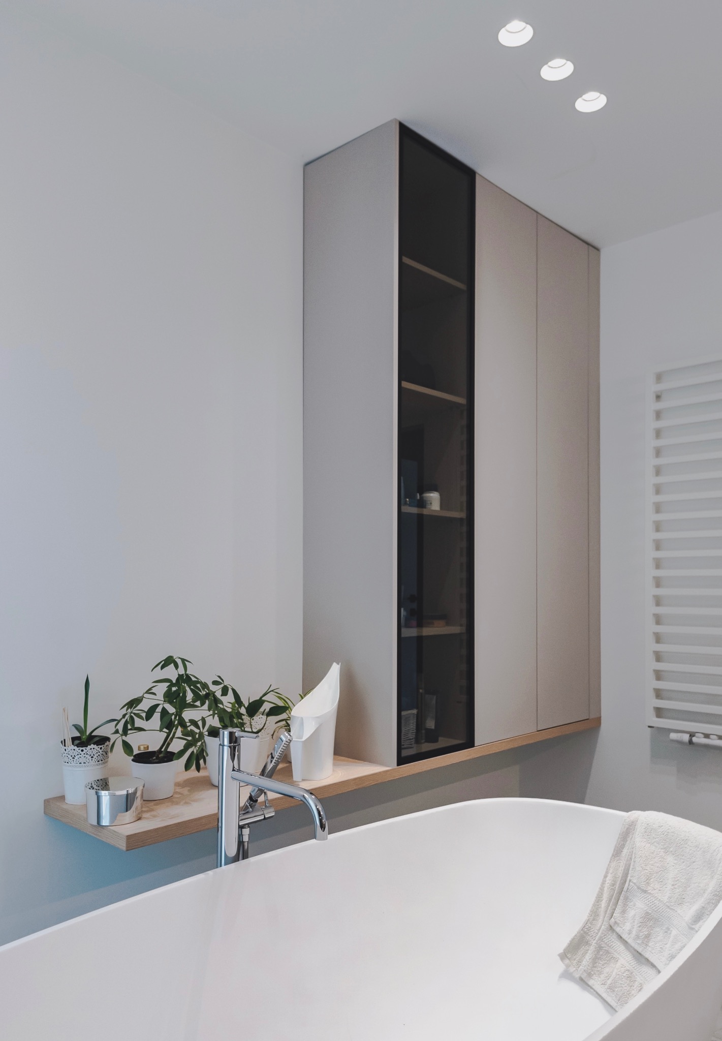 Jasna łazienka z wanną wolnostojącą, szafką wiszącą z przeszklonym frontem i drewnianą półką z roślinami doniczkowymi. Minimalistyczny design.