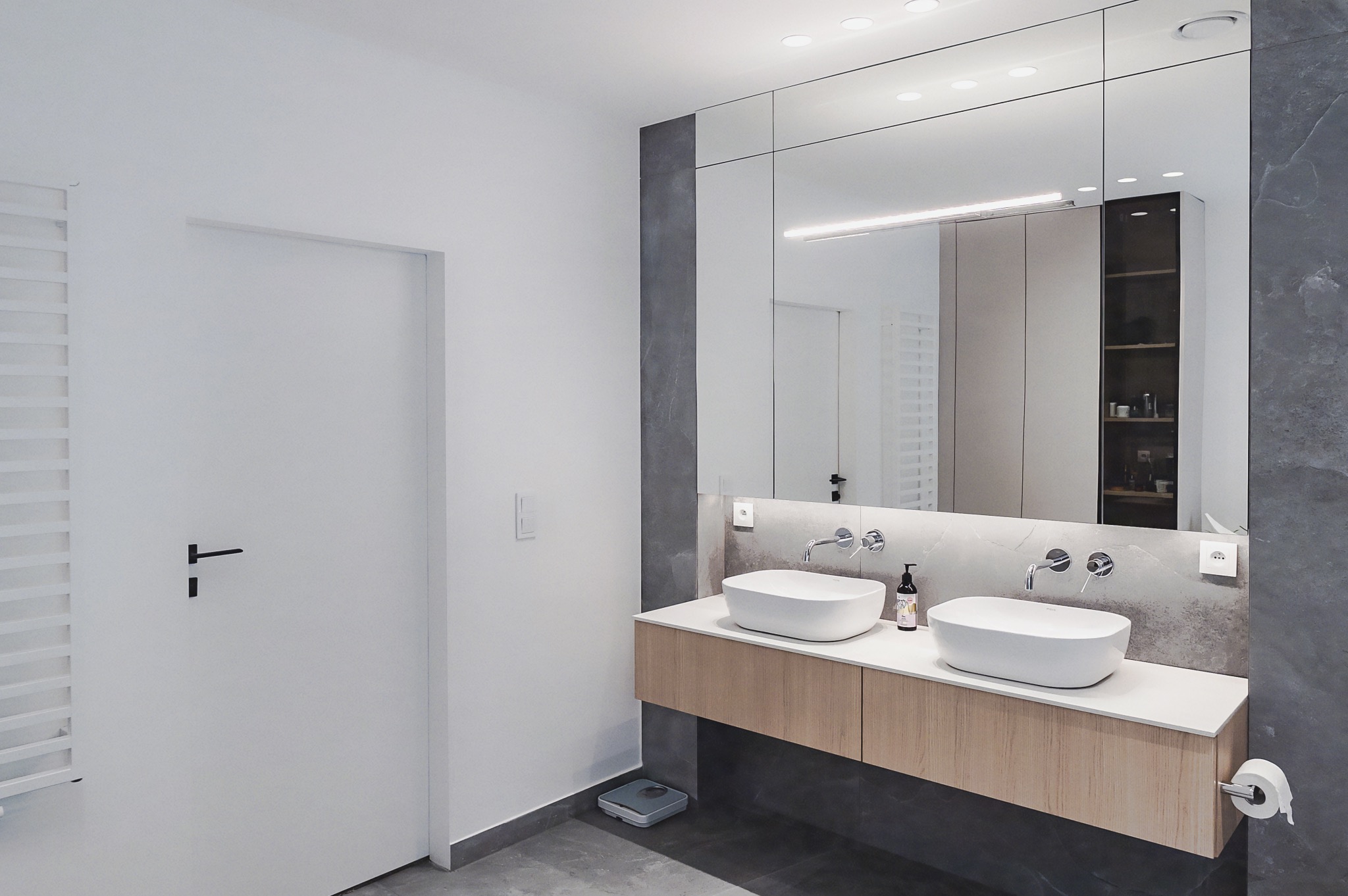 Nowoczesna łazienka z dwoma białymi umywalkami na drewnianej szafce, duże lustro z podświetleniem, szara ściana i podłoga, białe drzwi i grzejnik.