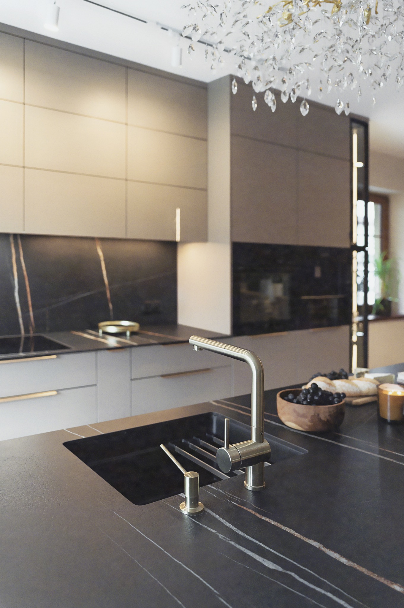 Nowoczesna kuchnia z czarnym blatem z żyłami, złotą armaturą i kryształowym żyrandolem. Minimalistyczne fronty szafek i oświetlenie LED dodają elegancji.