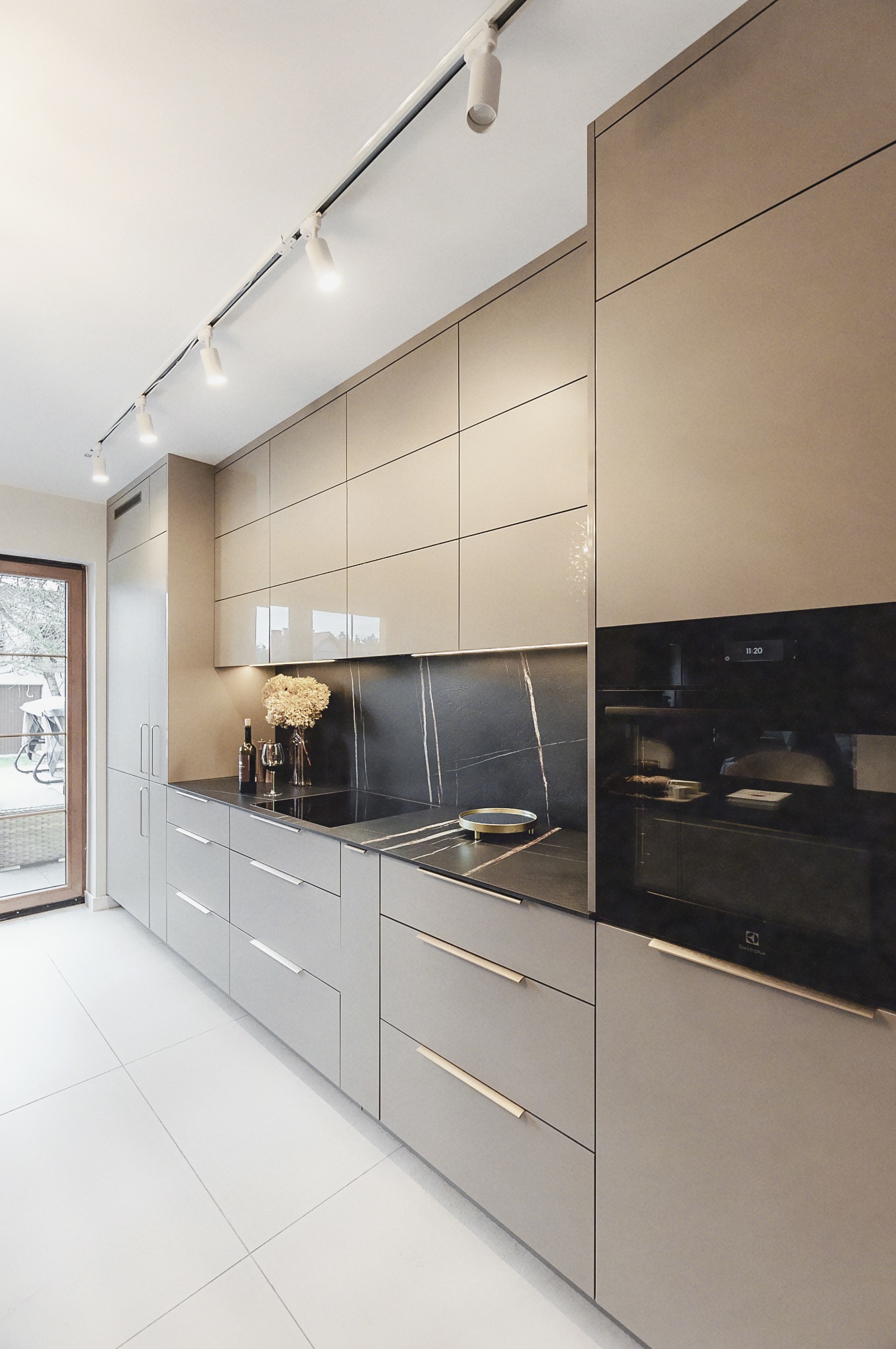 Nowoczesna kuchnia z szarymi frontami, czarnym blatem z marmurowym wzorem i złotymi uchwytami. Oświetlenie szynowe i widok na taras przez przeszklone drzwi.