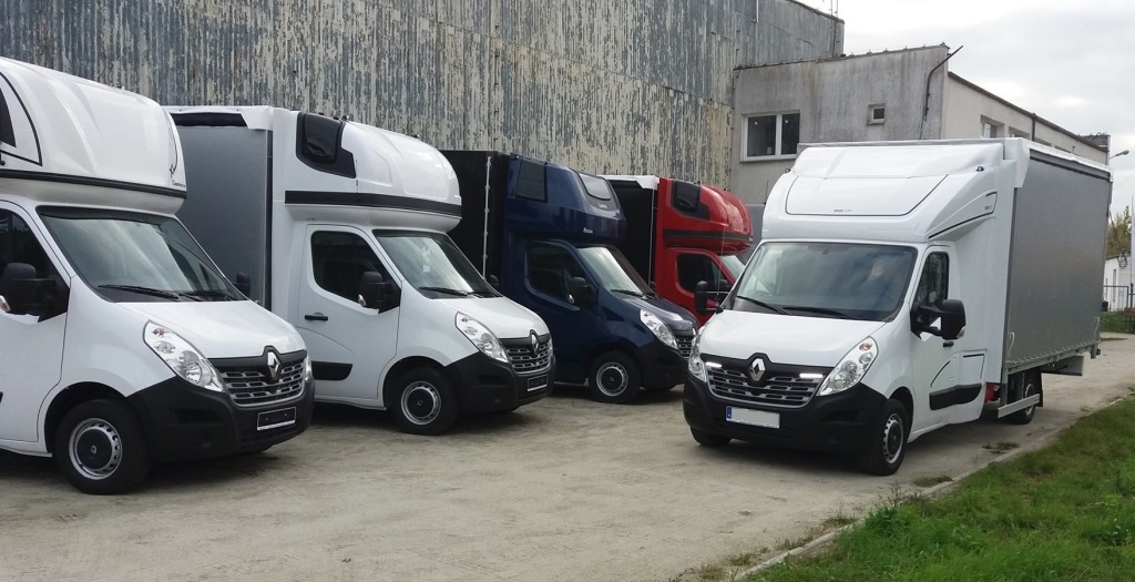 Flota samochodów dostawczych Renault Master w kolorach białym, granatowym i czerwonym, zaparkowana przed budynkiem z obdrapaną elewacją.