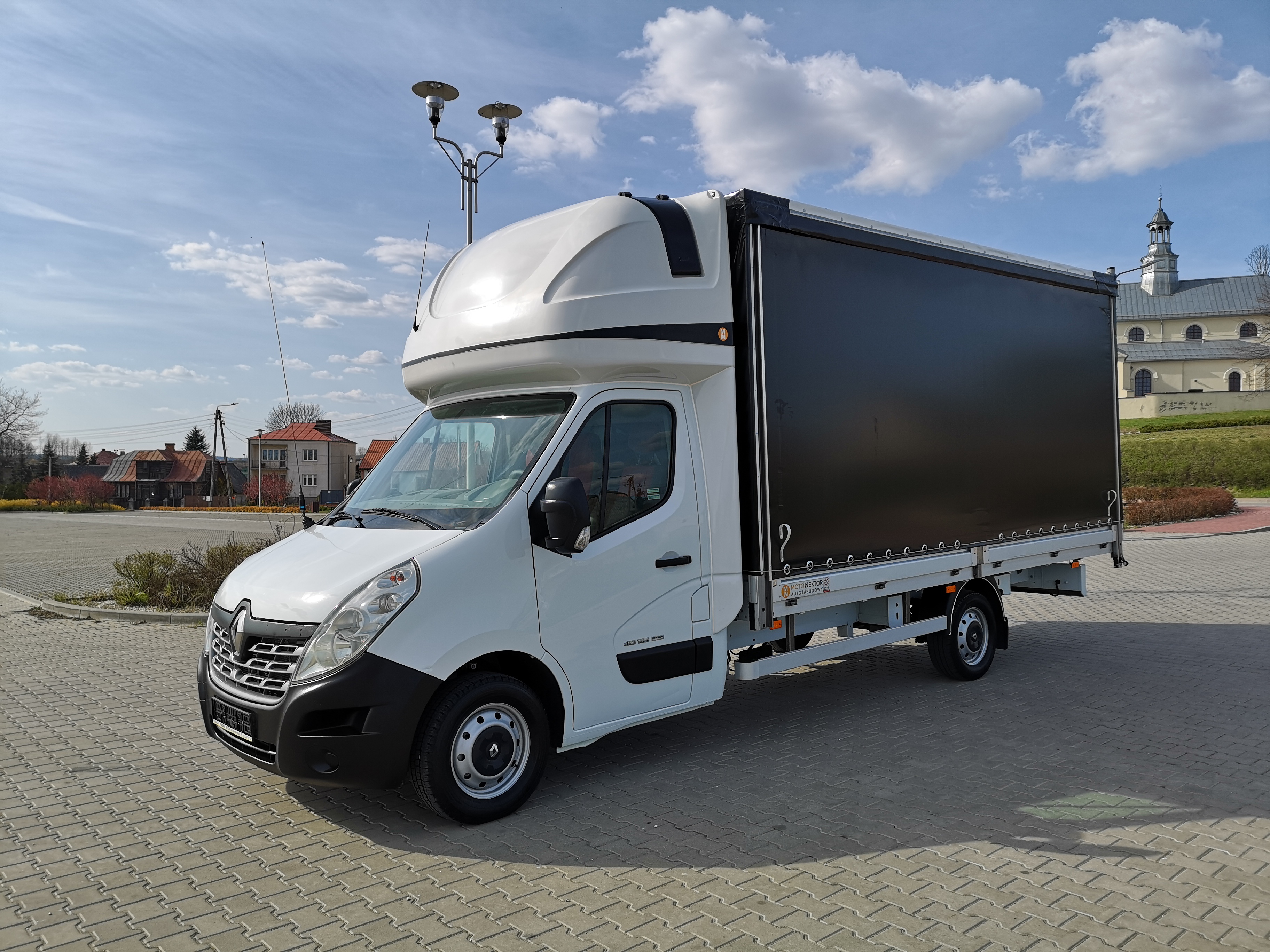 Biały Renault Master z czarną plandeką na tle brukowanego placu i kościoła w oddali, widok z boku.