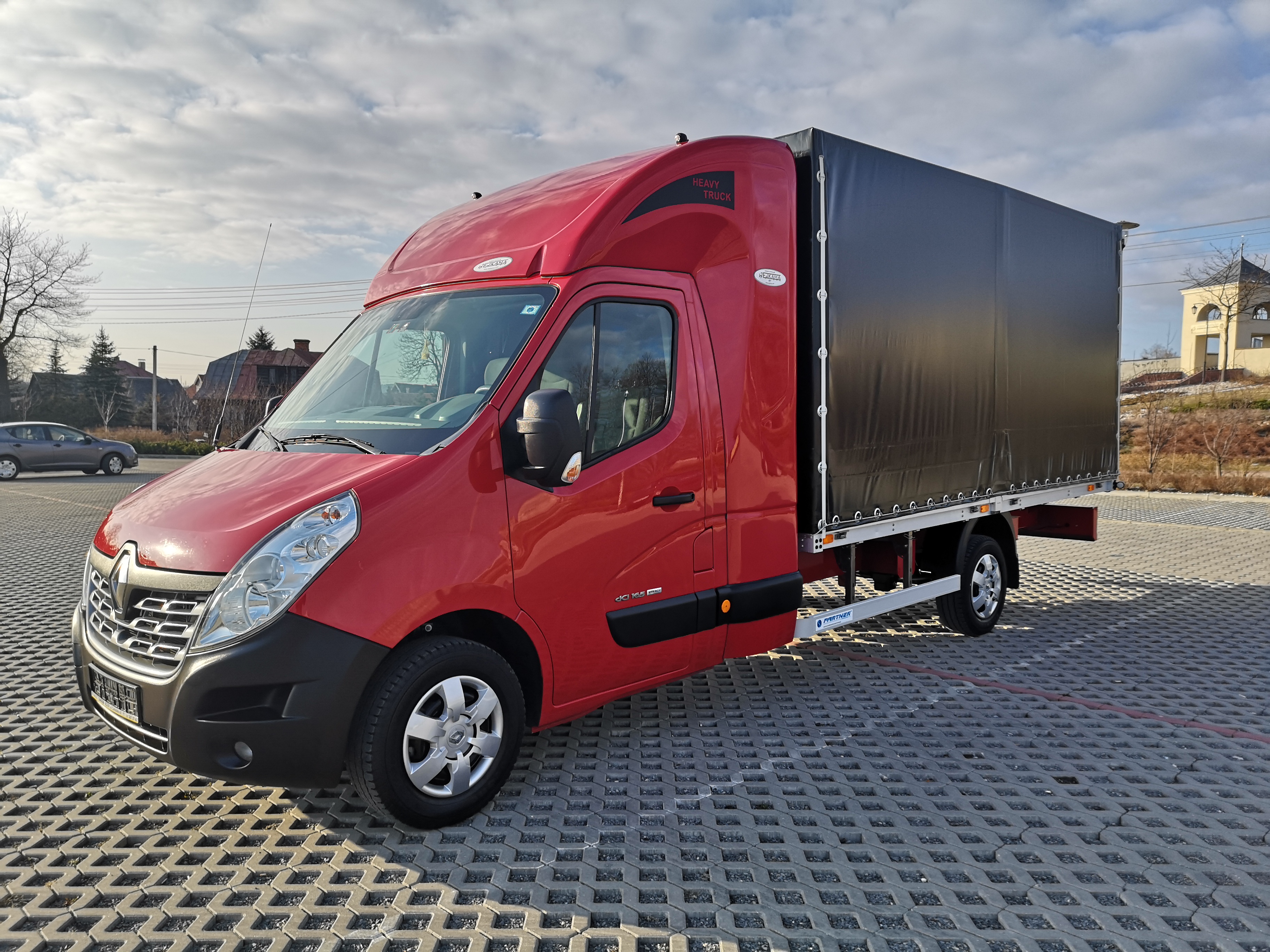 Czerwony bus z czarną plandeką zaparkowany na betonowej kostce, widoczny z boku, na tle budynków i pochmurnego nieba.