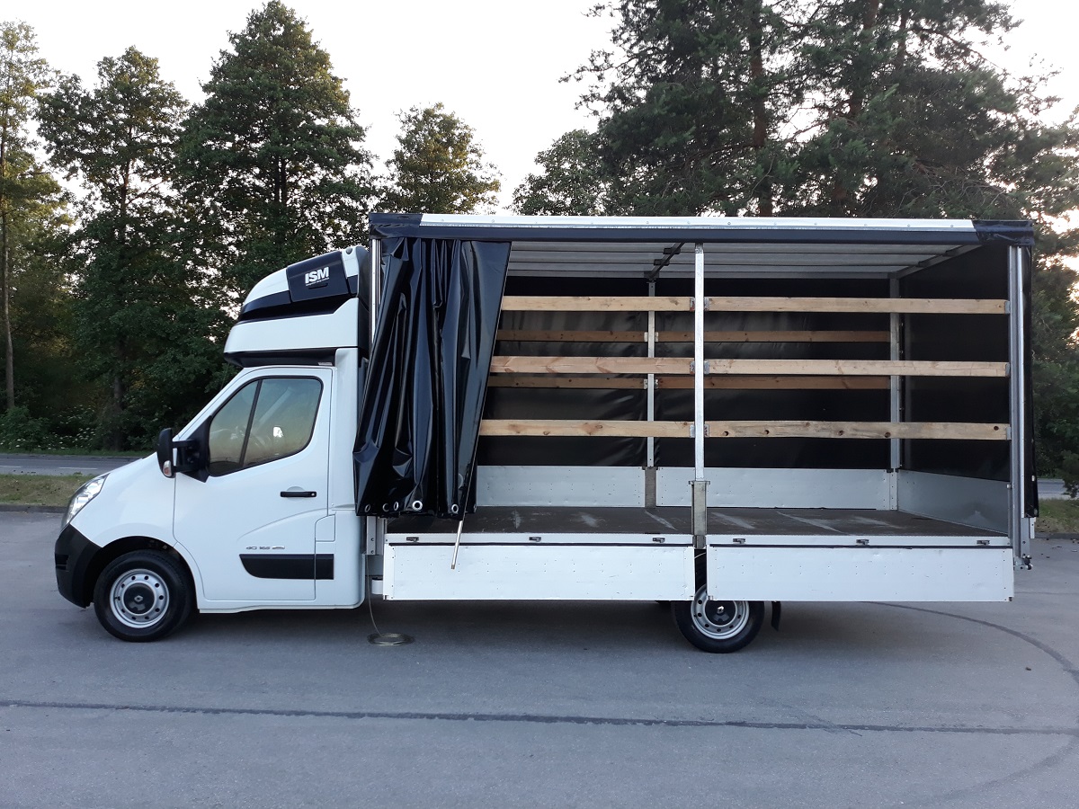 Biały samochód dostawczy z otwartą przestrzenią ładunkową, zabezpieczoną czarną plandeką i drewnianymi listwami, widok z boku na tle zieleni.