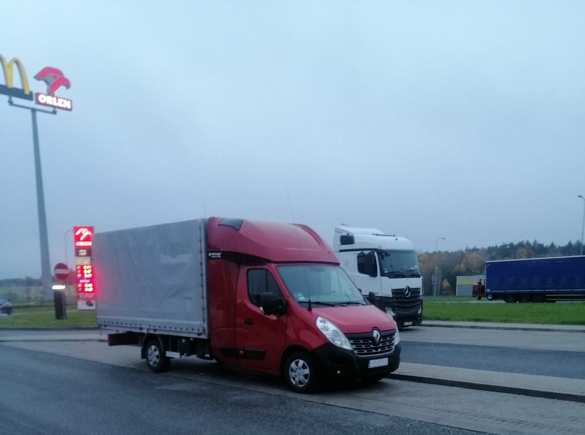 Czerwony bus z plandeką marki Renault zaparkowany na stacji benzynowej Orlen, w tle biała ciężarówka i zalesiony krajobraz w pochmurny dzień.