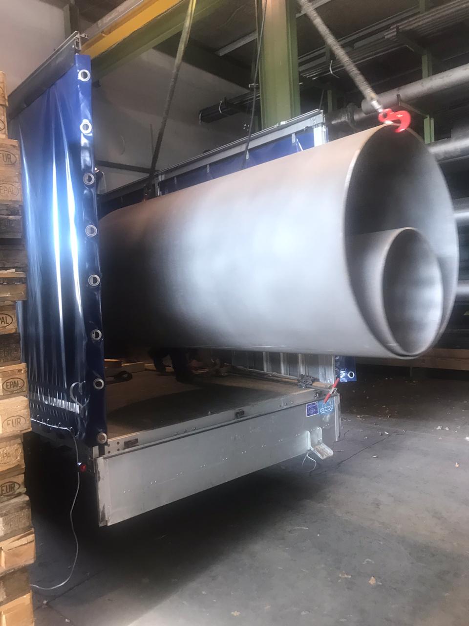 Załadunek dwóch dużych, srebrnych rur o różnych średnicach do naczepy ciężarówki z niebieską plandeką, widoczne palety EUR w tle.