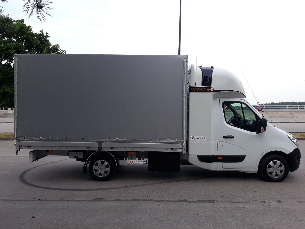 Biały samochód dostawczy z szarą plandeką na parkingu, widok z boku, zarys osoby na rowerze w tle.