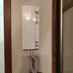 Biały panel domofonowy z szarą słuchawką spiralną zamontowany na beżowej ścianie obok brązowej futryny, widoczny również brązowy włącznik światła pod panelem.