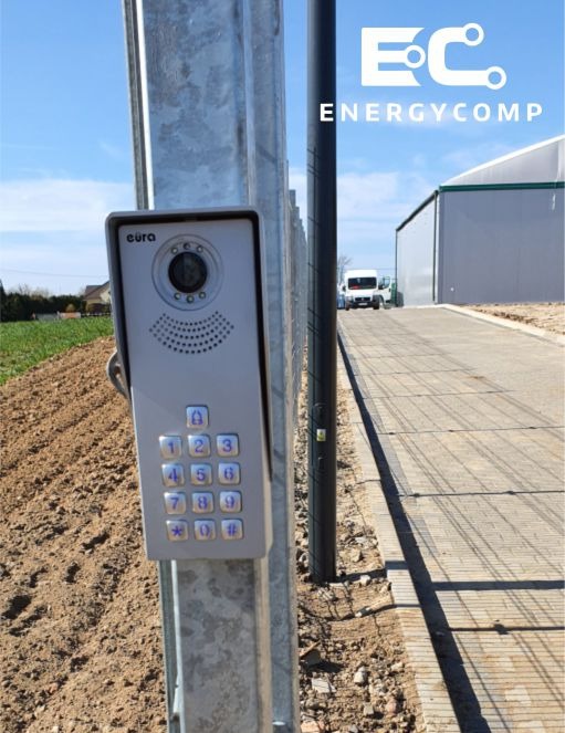 Szary wideodomofon Eura z klawiaturą numeryczną zamontowany na ocynkowanym słupku, widok z boku na tle placu budowy i błękitnego nieba.