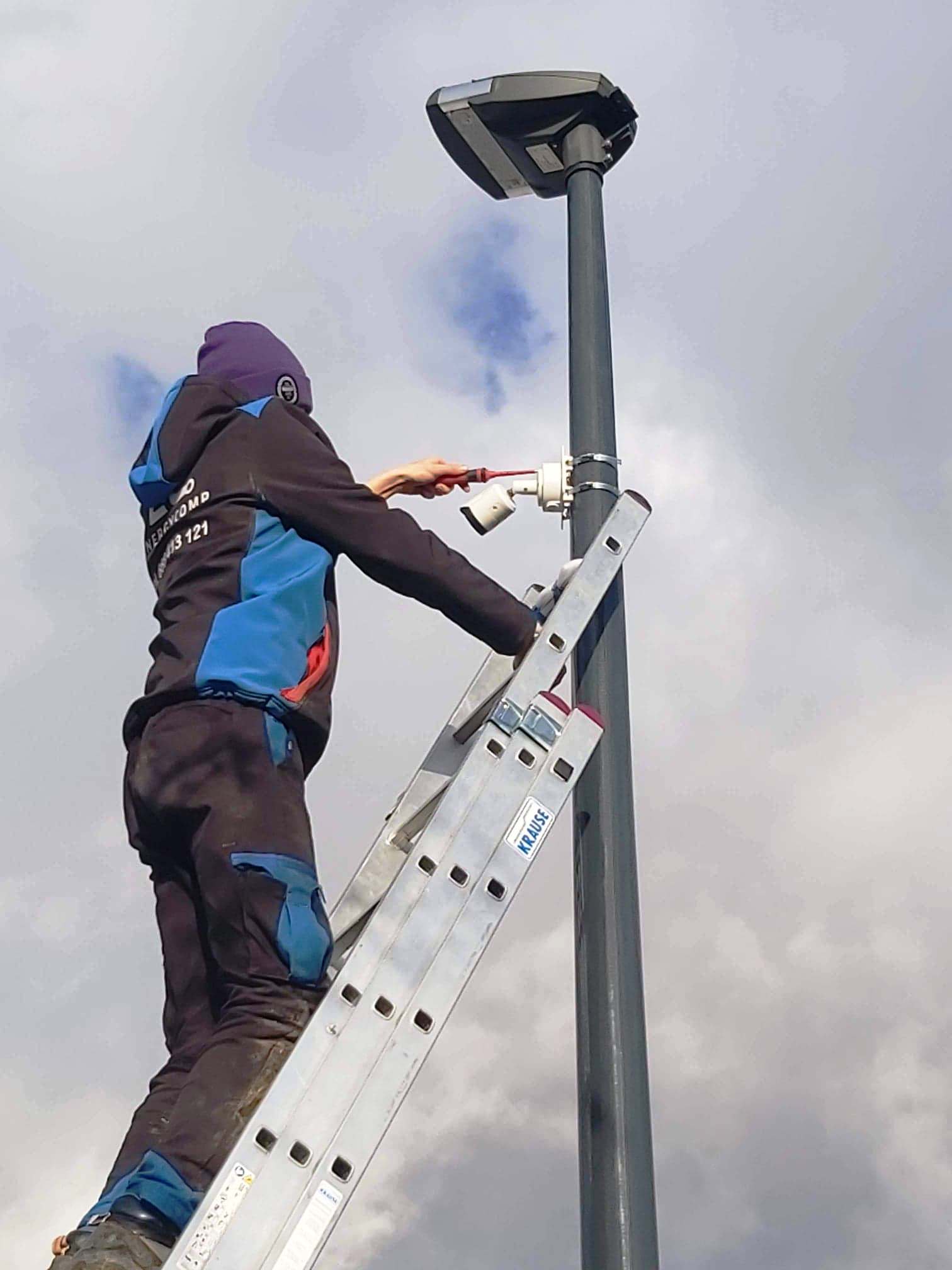 Instalator na drabinie mocuje kamerę monitoringu do słupa oświetleniowego na tle pochmurnego nieba.