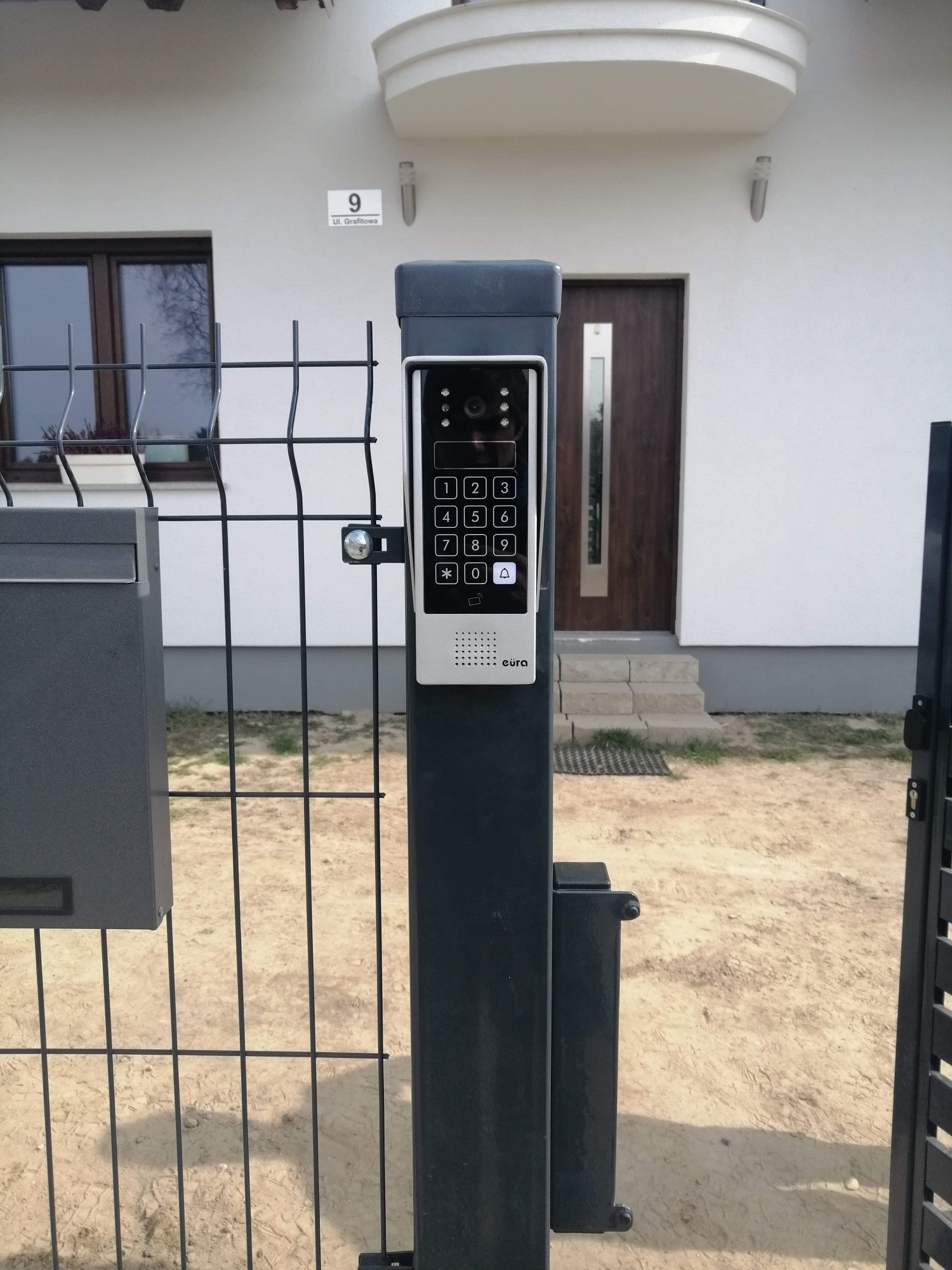Zamontowany wideodomofon Eura na metalowym słupku ogrodzenia posesji z widoczną skrzynką na listy i numerem domu na białej fasadzie budynku.