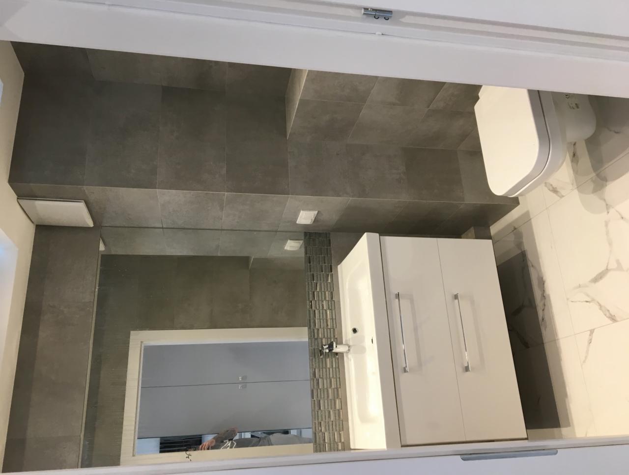 Wykończona łazienka z szarymi płytkami na ścianach i podłodze, białą szafką z umywalką i toaletą, marmurową ścianą oraz lustrem z mozaiką.