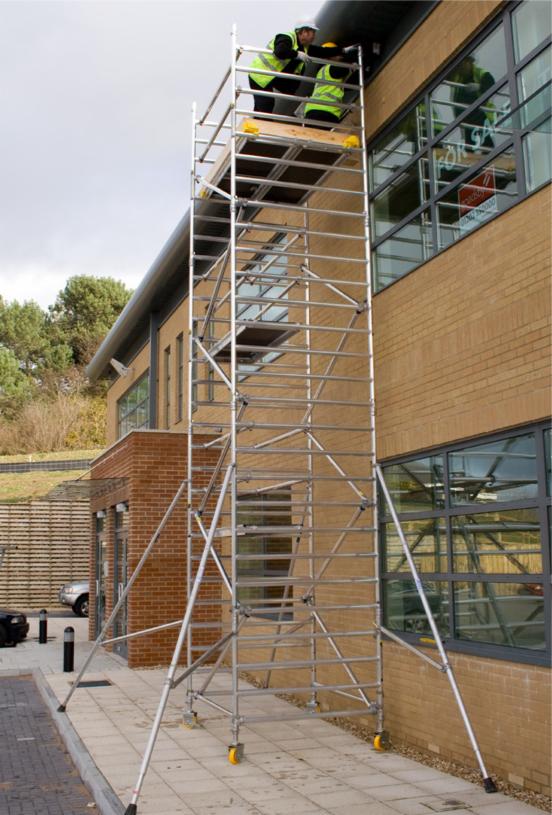Dwóch pracowników w kamizelkach odblaskowych na wysokim, metalowym rusztowaniu na kółkach przy ścianie budynku z cegły z dużymi oknami, na których widać odbicie otoczenia.