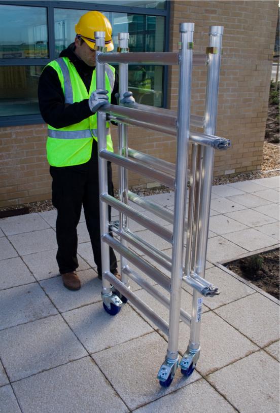 Pracownik w kasku i kamizelce odblaskowej rozkłada aluminiowe rusztowanie na kółkach przed budynkiem z cegły.