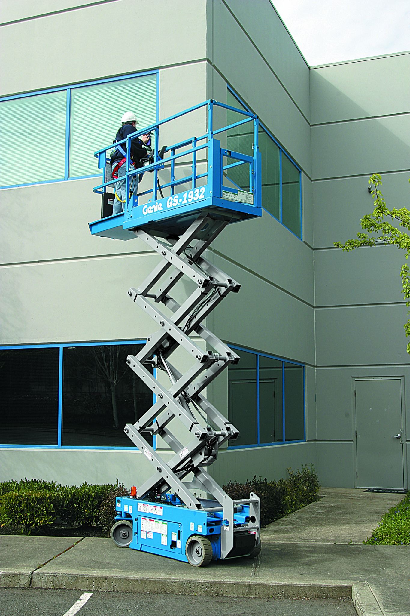 Pracownik w kasku i uprzęży na podnośniku nożycowym Genie GS-1932, wykonujący prace na wysokości przy szarym budynku biurowym z dużymi oknami.