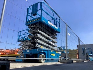 Niebieski podnośnik nożycowy na placu budowy, zabezpieczony siatką ogrodzeniową, widoczny w słoneczny dzień.