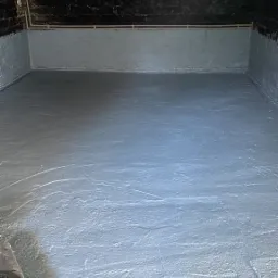 Świeżo pomalowane pomieszczenie gospodarcze z szarą podłogą i ścianami, widoczne ślady pędzla, surowy mur z czarnym pasem u góry, sufit betonowy, żarówka na kablu.