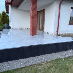 ABW - Wyłożony jasnoszarą płytką taras przy domu z ceglanym filarem. Widoczne czarne obrzeża i przygotowane podłoże pod dalsze prace wykończeniowe.