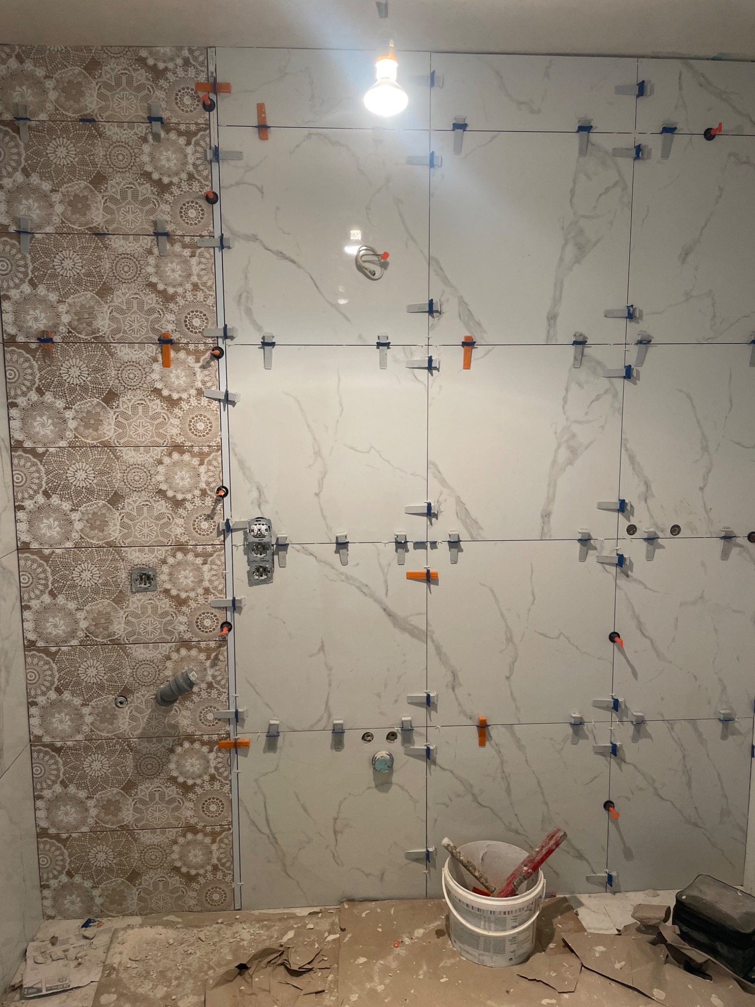 Układanie płytek w łazience: białe płytki imitujące marmur i dekoracyjne płytki z motywem koronki, krzyżyki dystansowe, widoczne narzędzia i wiadro z klejem.