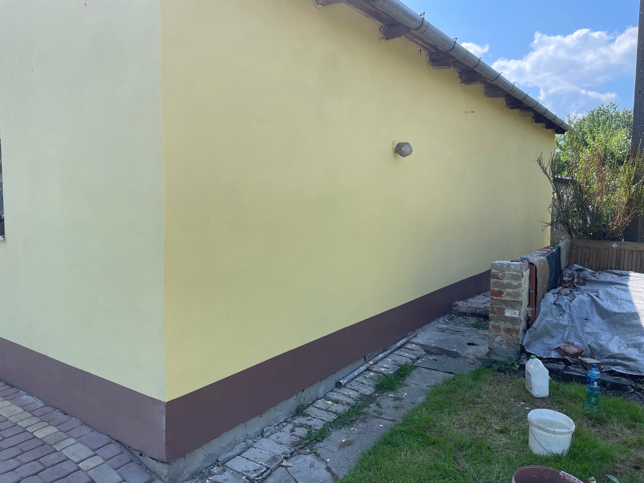 Żółta elewacja budynku po malowaniu z brązowym cokołem, widok z zewnątrz. Na trawie wiadro i butelki. Świeżo pomalowana ściana w słoneczny dzień.