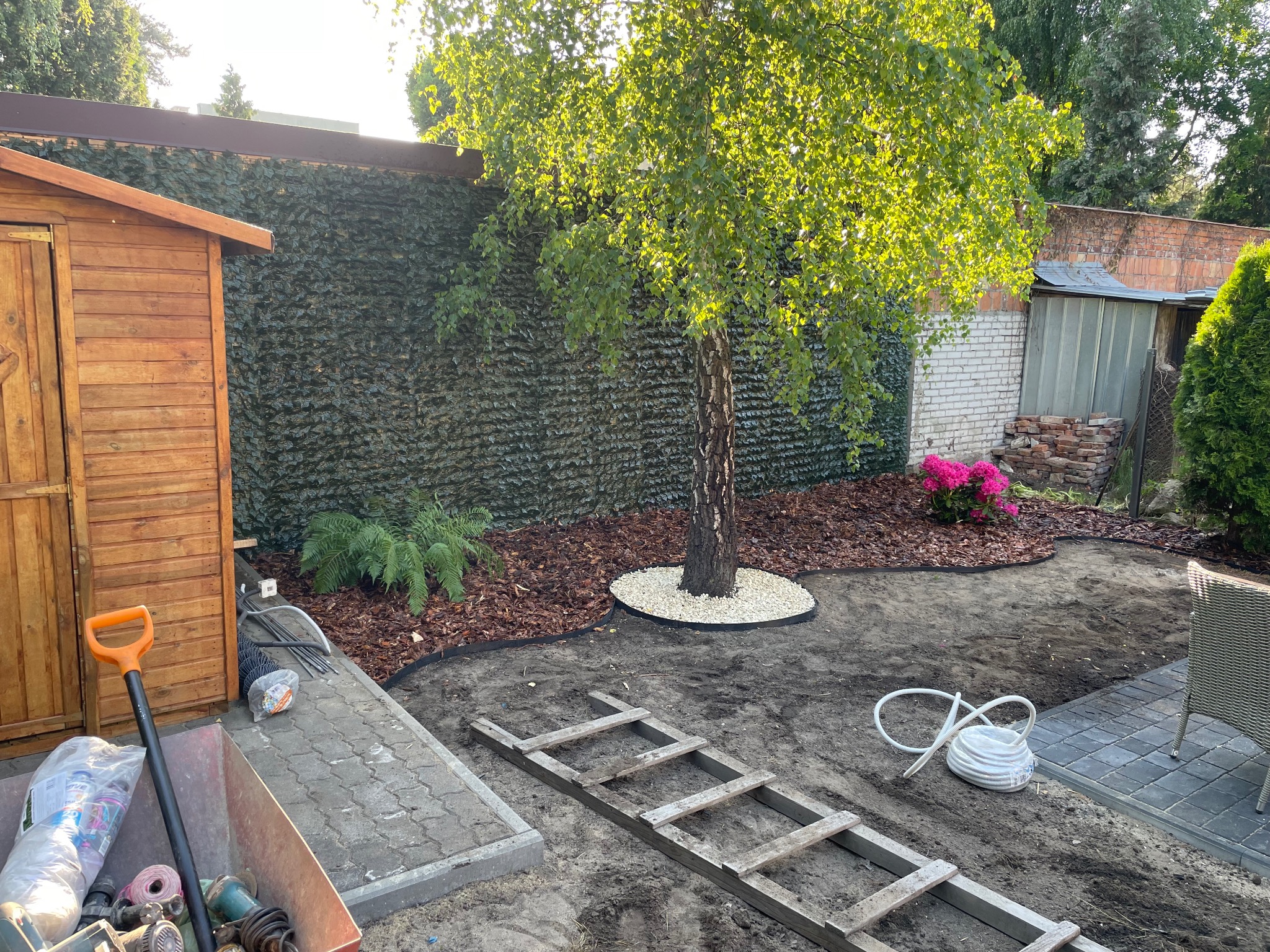 Aranżacja ogrodu: drzewo otoczone białymi kamieniami, kora, trawa, drewniana altana, narzędzia ogrodnicze i drabina. Tło stanowi zielona ściana z roślin.