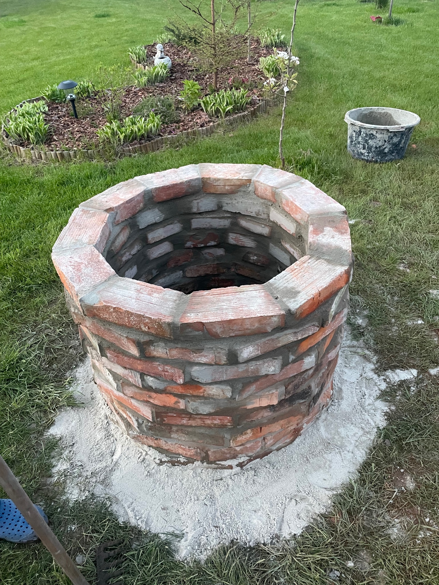Murowana studnia z czerwonej cegły na trawniku, obok wiadro i rabata z roślinami. Widoczne ślady zaprawy. Nietypowe ujęcie z góry.