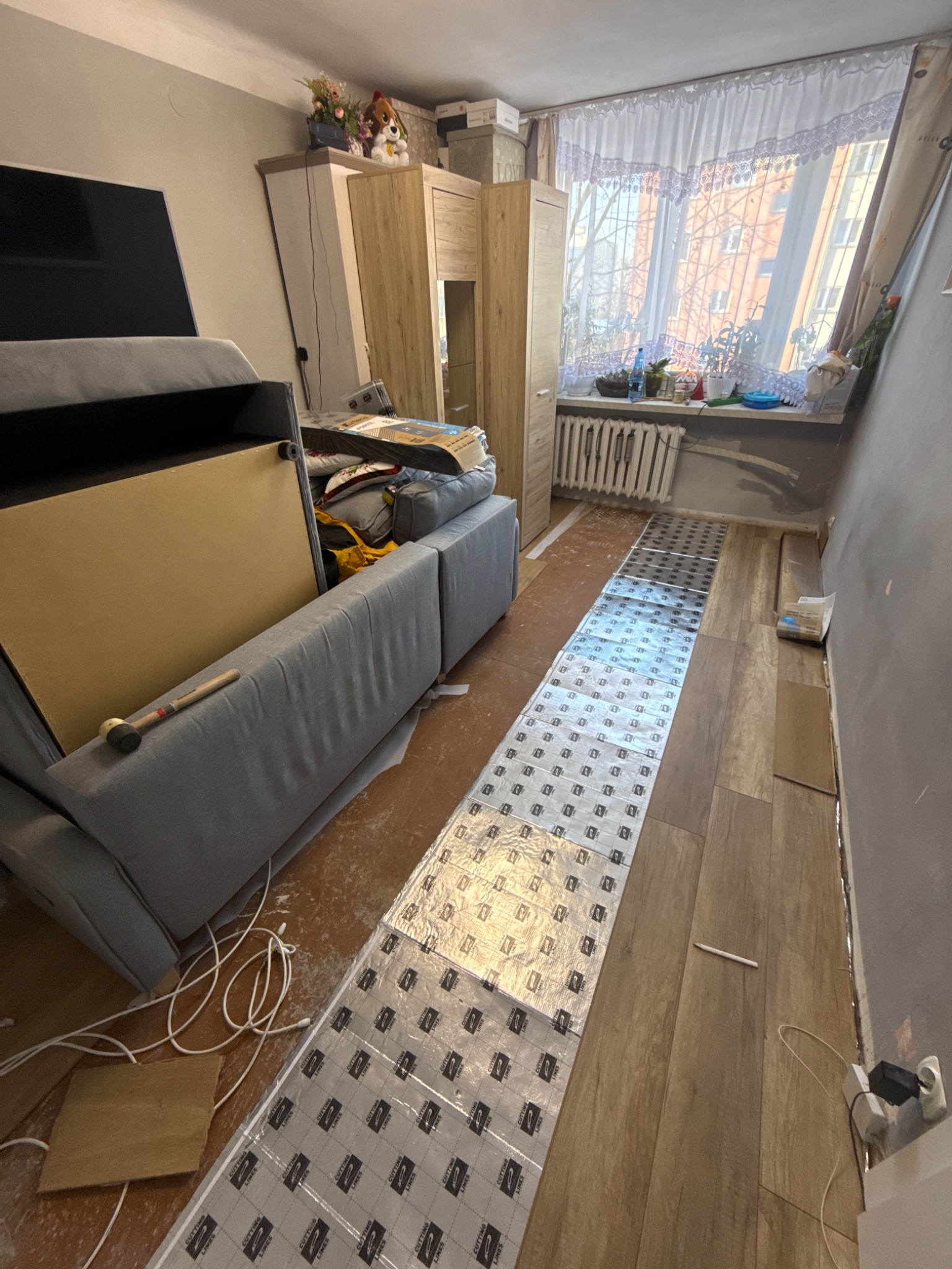 Układanie paneli podłogowych w pokoju z częściowo odsuniętym meblem. Widoczna folia izolacyjna i panele. Młotek leży na sofie. Okno z firankami w tle.