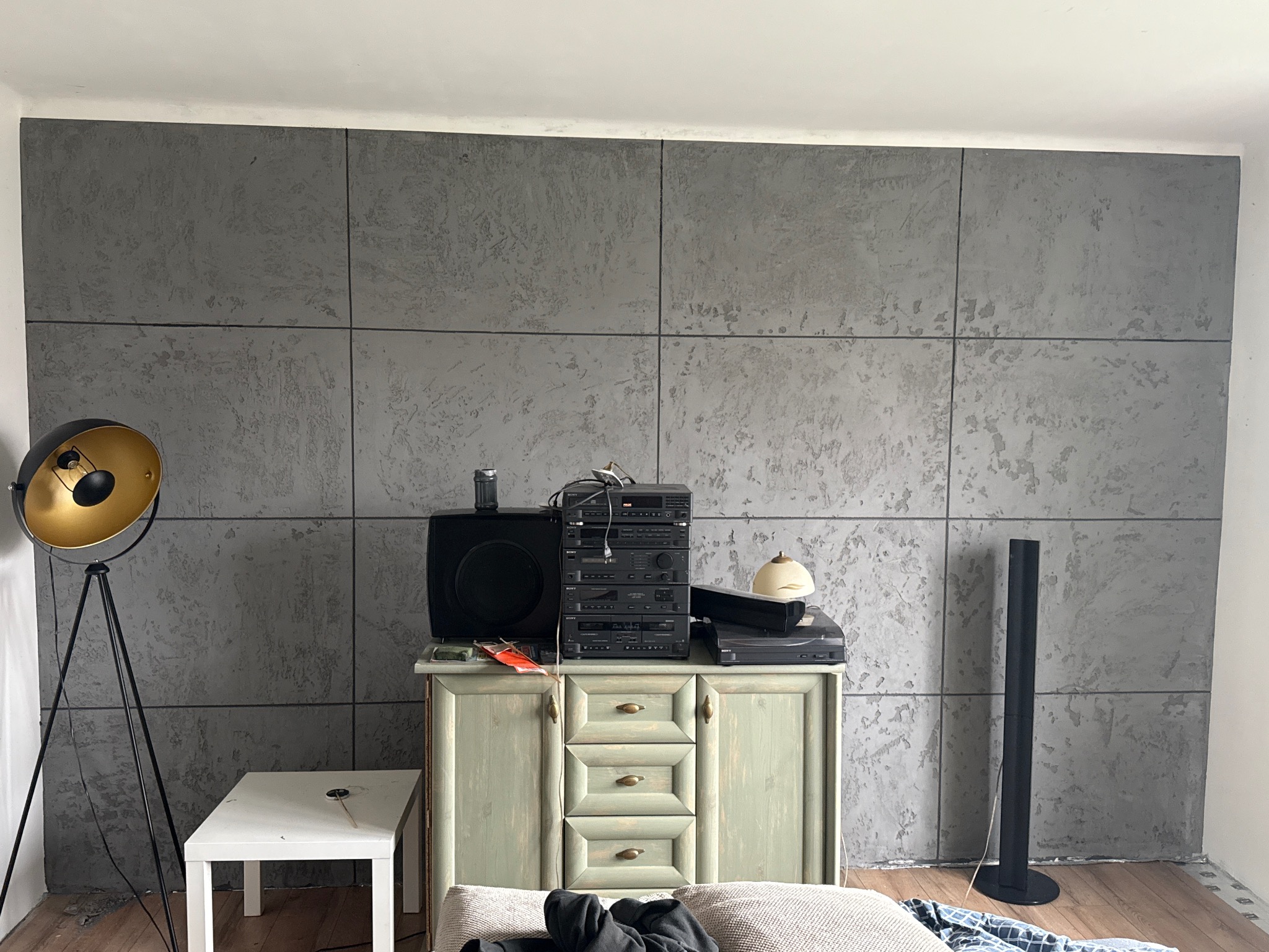 Ściana wykończona panelami imitującymi beton, z widocznym sprzętem audio na komodzie i lampą stojącą. Szare panele tworzą nowoczesny efekt w pomieszczeniu.
