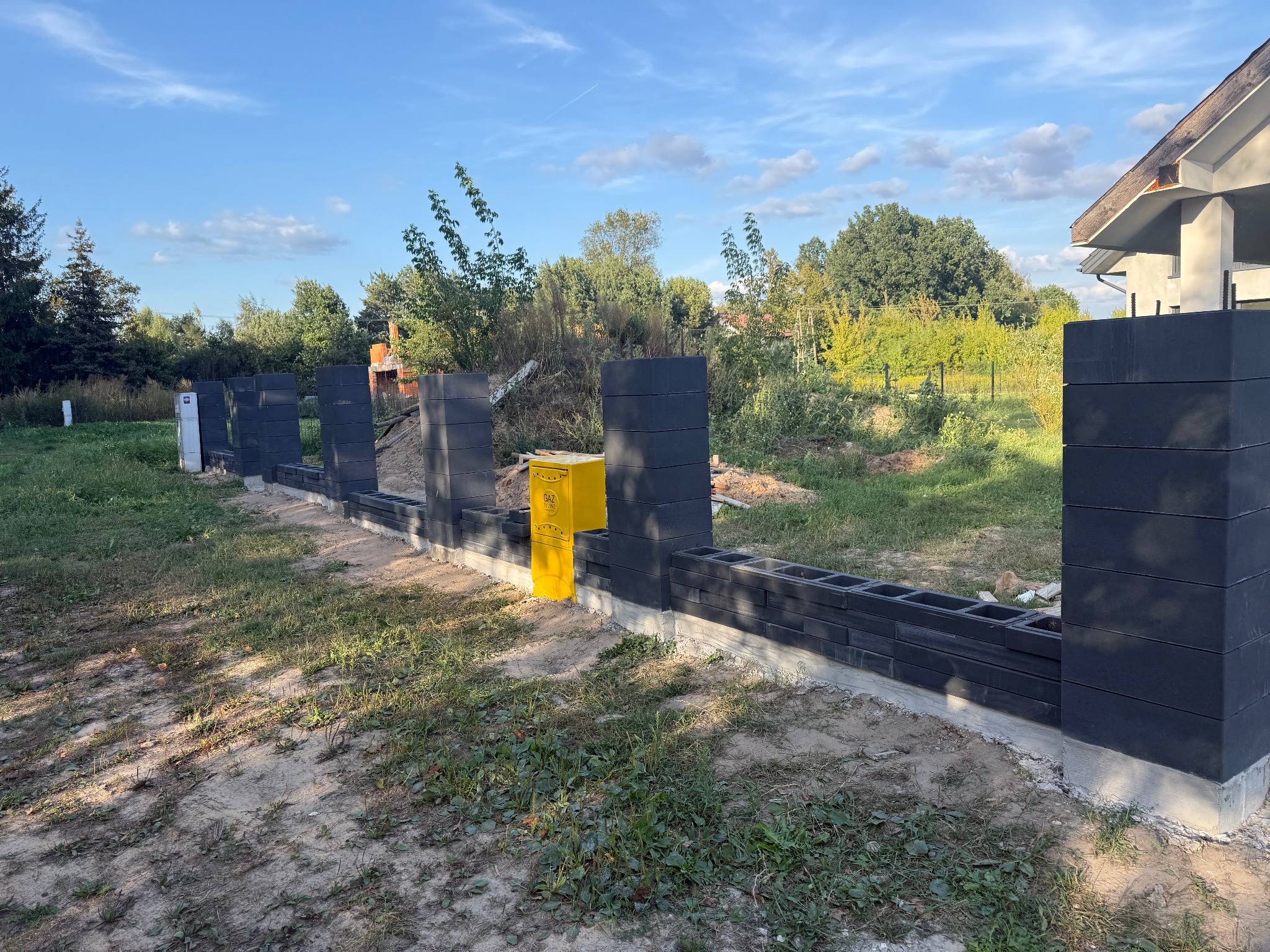 Wznoszenie ogrodzenia z szarych, betonowych bloczków na fundamencie. Żółta skrzynka gazowa przy jednym ze słupków. Widok na posesję i zieleń w tle, błękitne niebo.