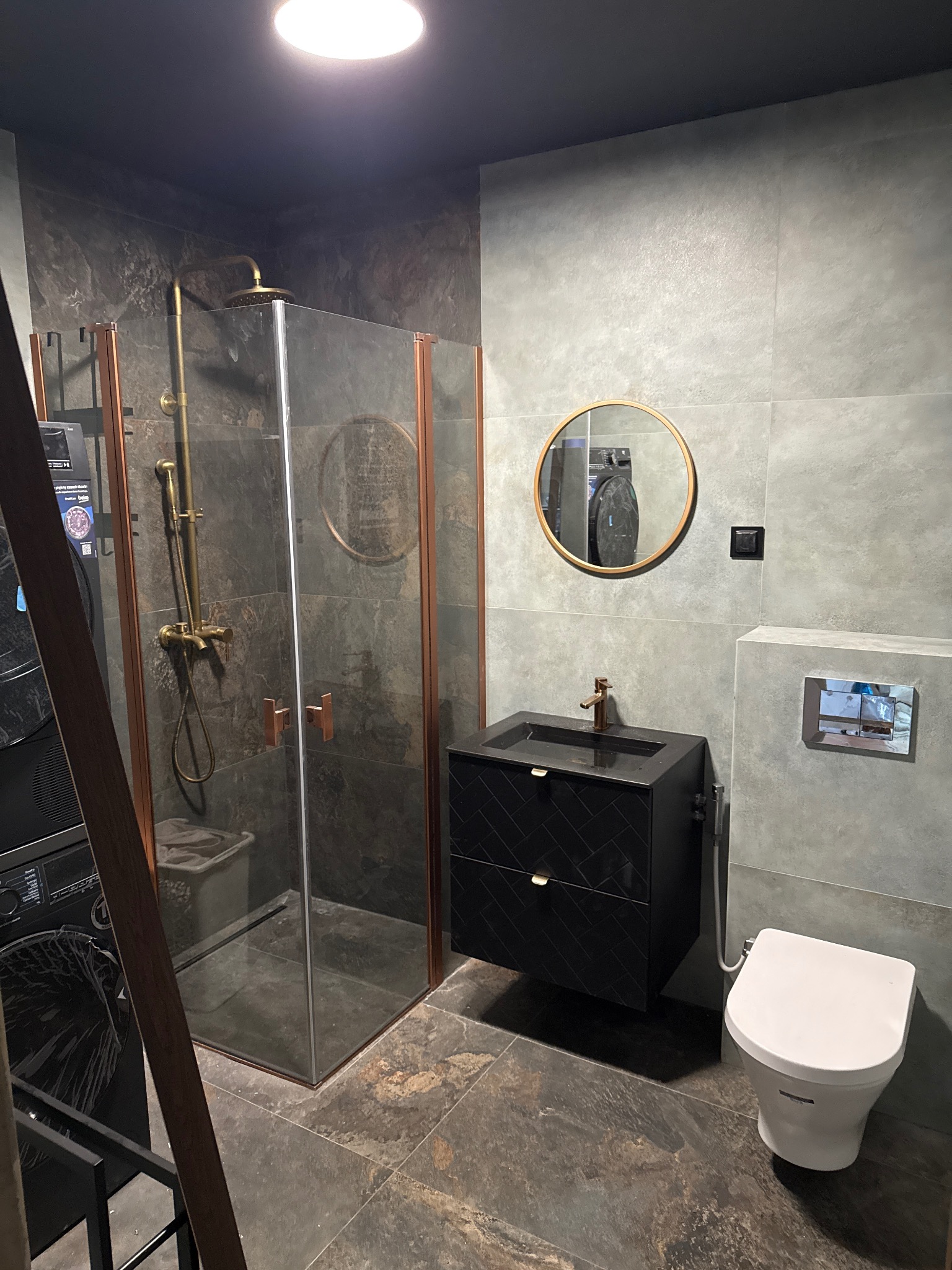 Nowoczesna łazienka z kabiną prysznicową w stylu loft, czarną szafką z umywalką i okrągłym lustrem. Szare płytki na ścianach i podłodze.