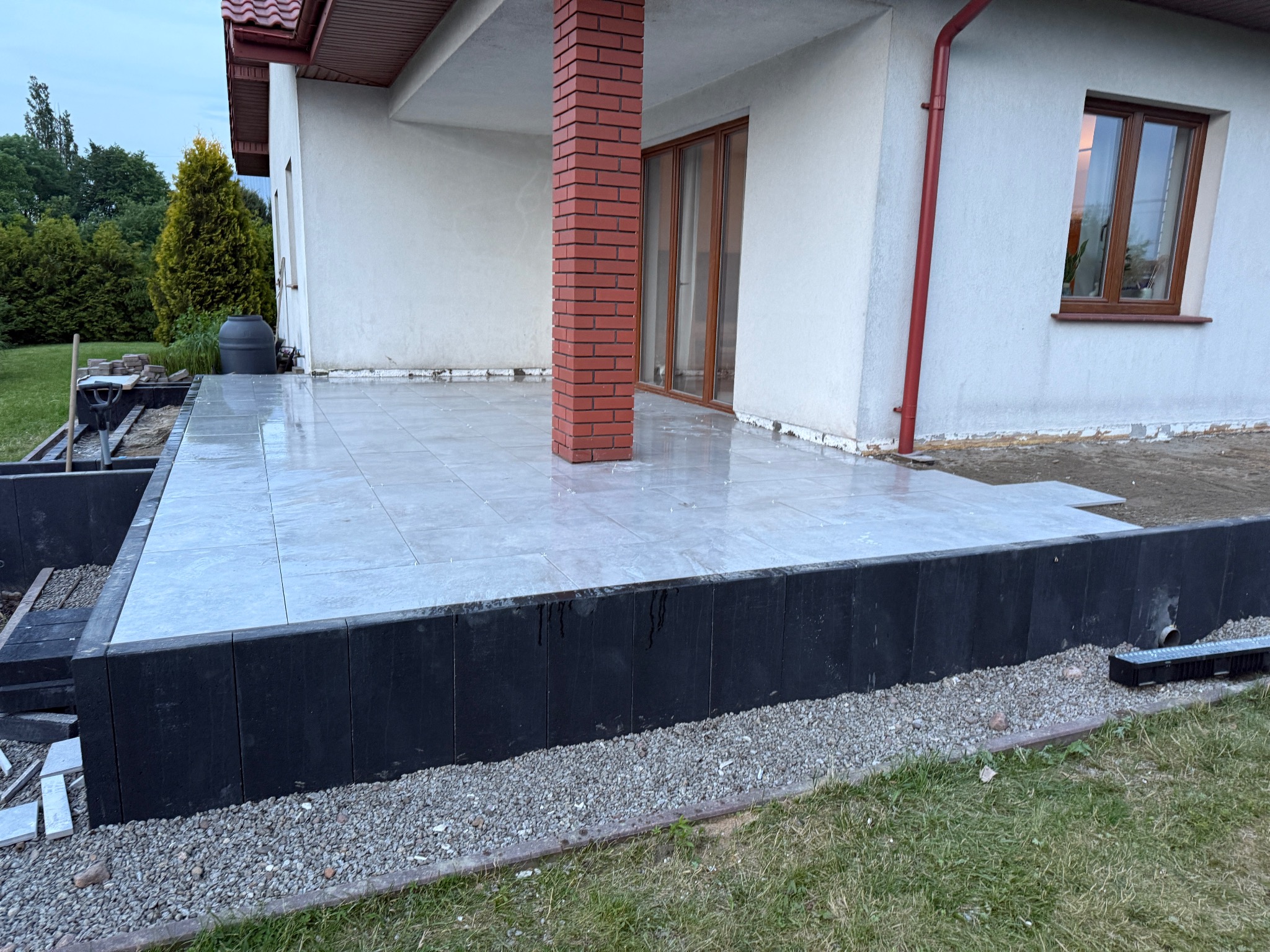 Wyłożony jasnoszarą płytką taras przy domu z ceglanym filarem. Widoczne czarne obrzeża i przygotowane podłoże pod dalsze prace wykończeniowe.