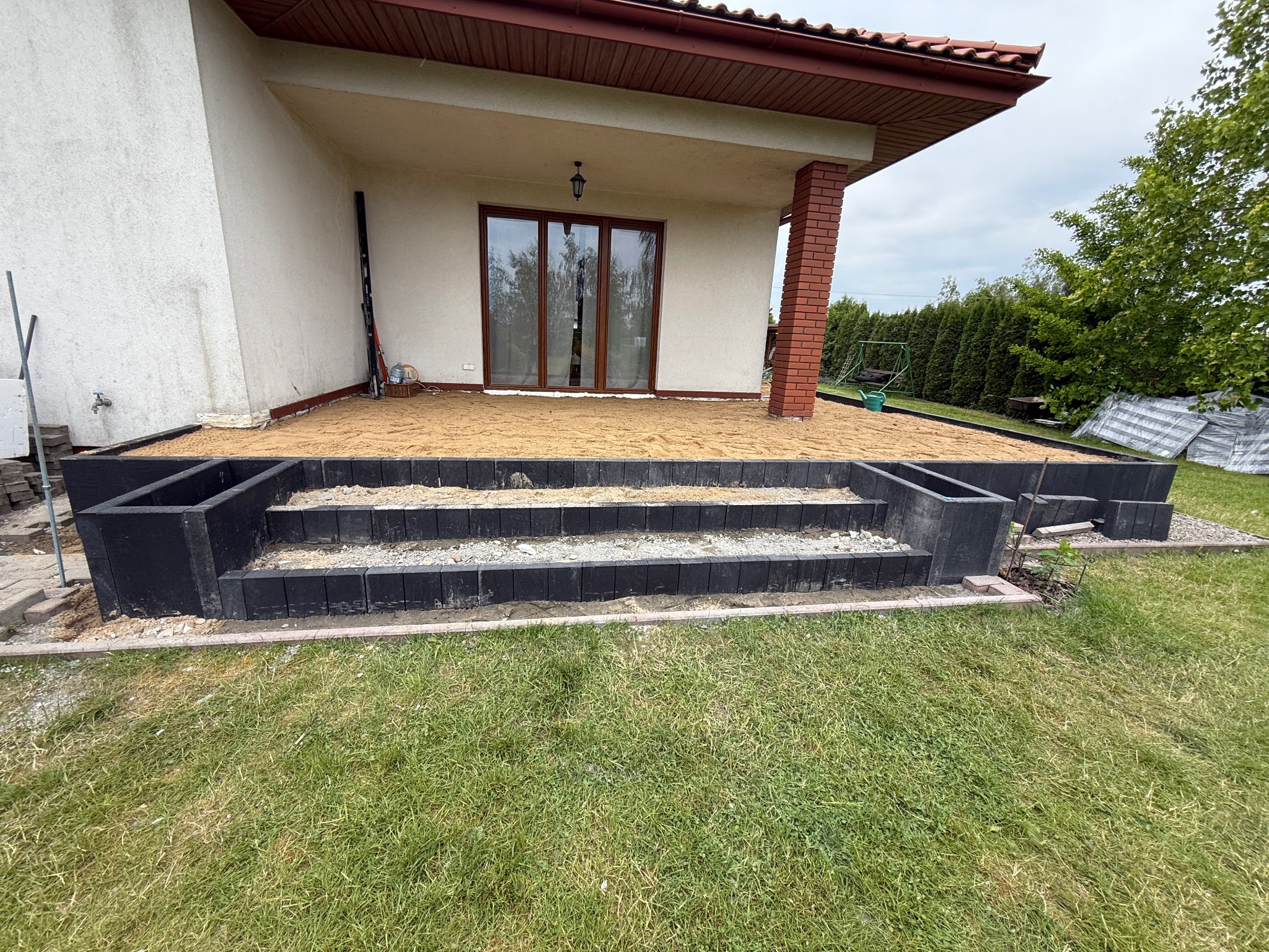 Budowa tarasu z kostki brukowej: czarne obrzeża, piasek, widok na dom z ceglaną kolumną i zielony ogród. Etap przygotowania podłoża.