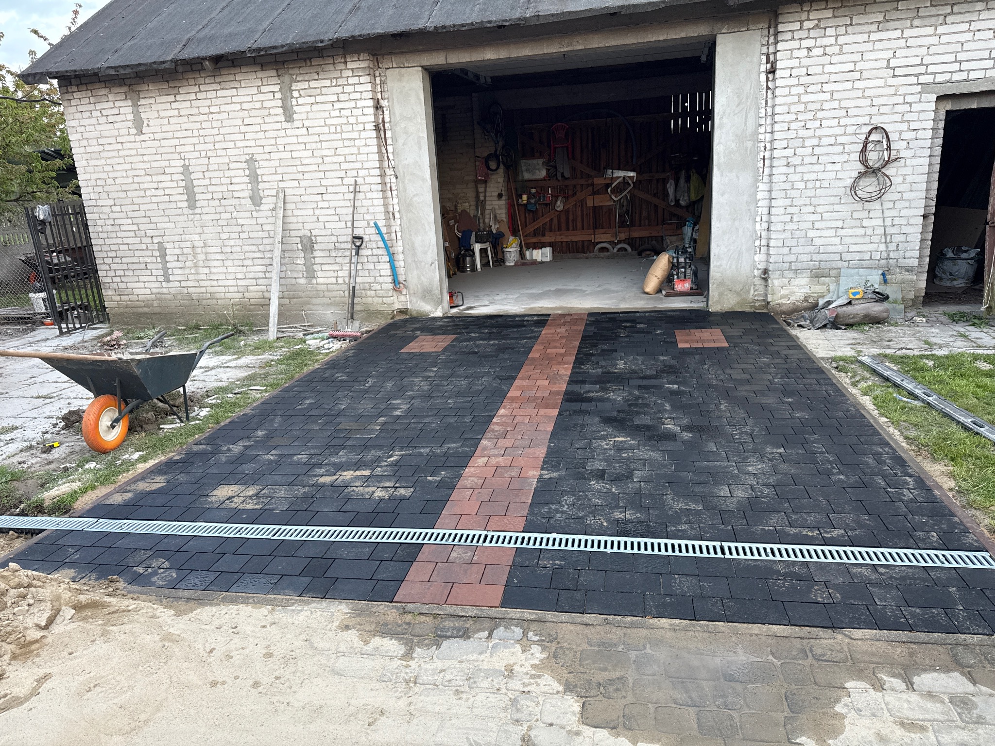 Ułożona kostka brukowa w kolorze czarnym i czerwonym z odpływem liniowym przed garażem. Widoczny wózek i narzędzia.