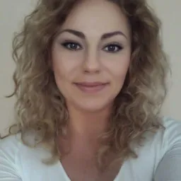 Portret kobiety z blond, kręconymi włosami w białej bluzce na jednolitym tle.