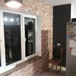 Ściana z cegły dekoracyjnej w mieszkaniu, widoczne okno z białymi ramami, czarna ściana obok, krzesło i lodówka w tle.