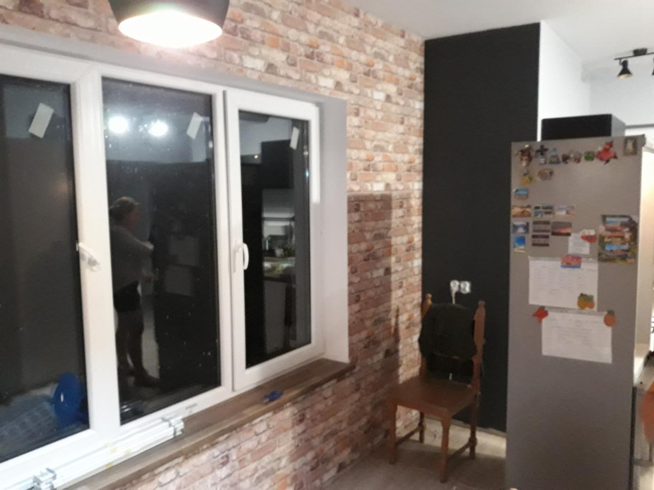 Ściana z cegły dekoracyjnej w mieszkaniu, widoczne okno z białymi ramami, czarna ściana obok, krzesło i lodówka w tle.