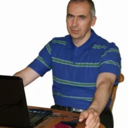 Mężczyzna w niebieskiej koszulce w paski siedzi przy drewnianym stole z laptopem i zewnętrznym dyskiem twardym, spoglądając w obiektyw.