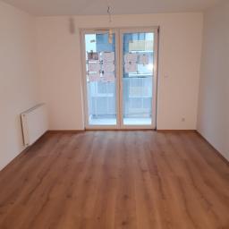 Progressfinishhome@gmail.com - Puste, białe pomieszczenie z panelami podłogowymi w odcieniach brązu, grzejnikiem na ścianie i planami mieszkania przyklejonymi do szklanych drzwi balkonowych.