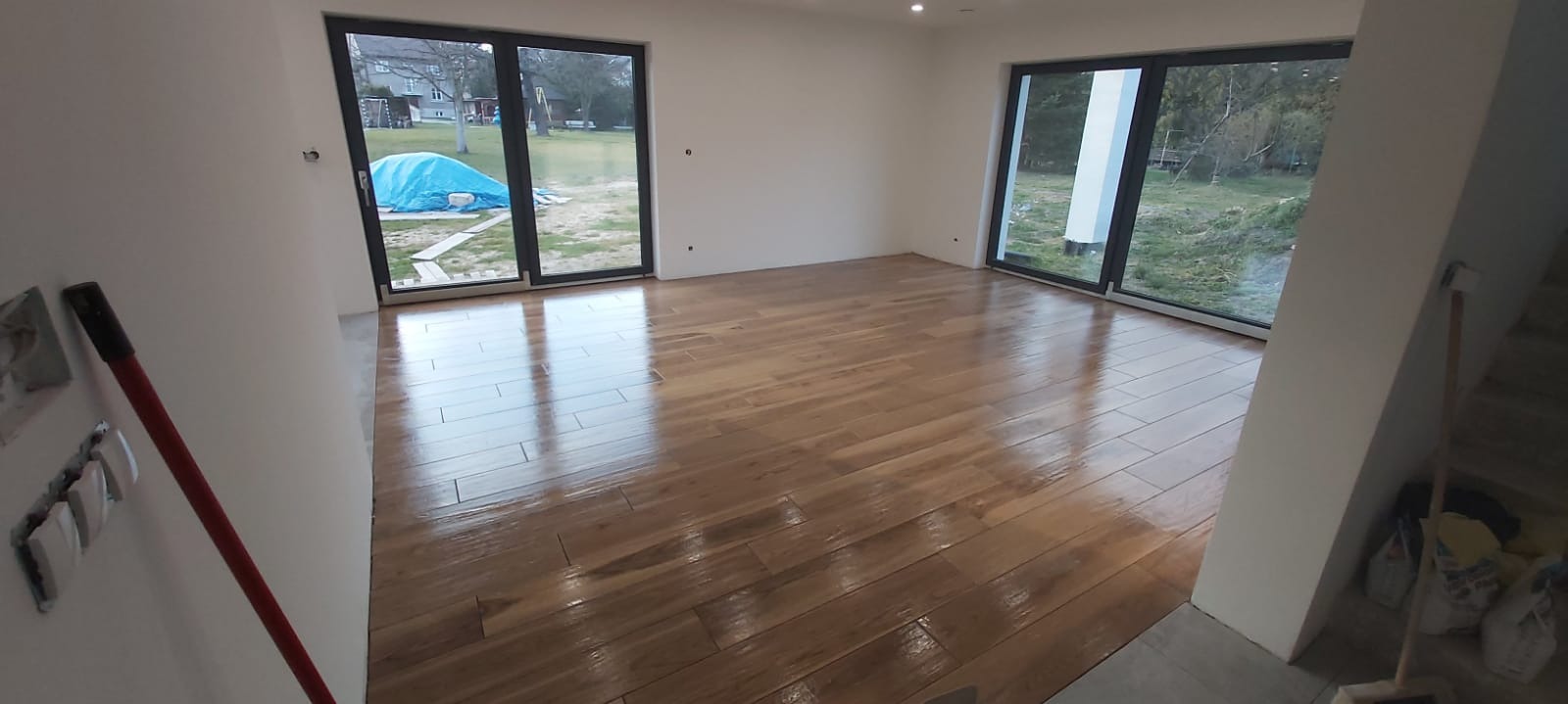 Jasny parkiet ułożony w jodełkę w nowym, przestronnym wnętrzu z dużymi oknami, widoczne przygotowania do wykończenia ścian i instalacji elektrycznej.
