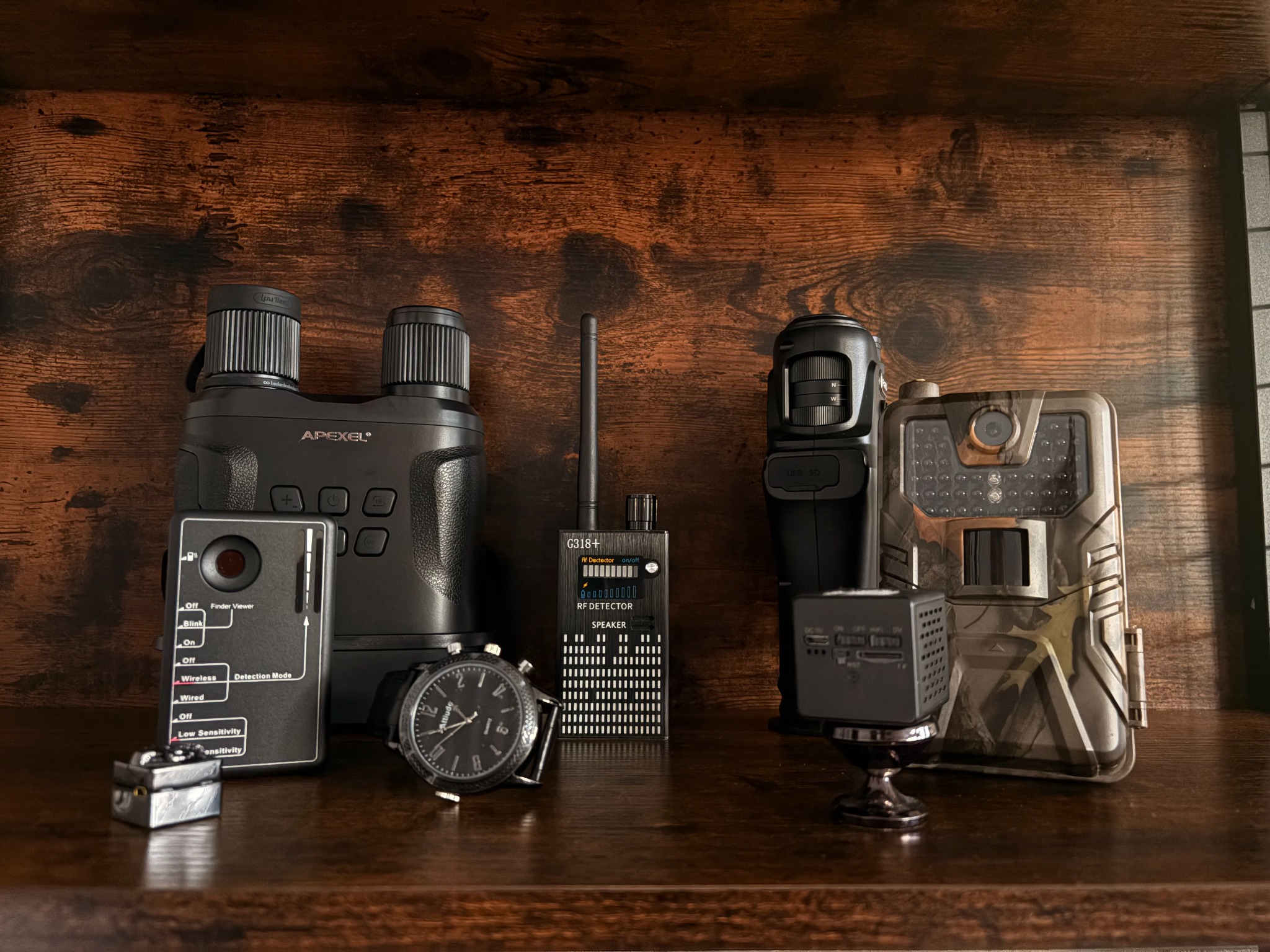 Zestaw detektywistyczny: lornetka noktowizyjna Apexel, detektor RF G318+, kamera szpiegowska, zegarek Altitude. Sprzęt na drewnianej półce.