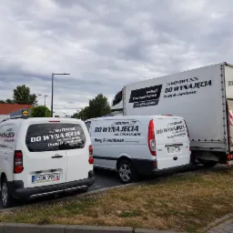 Flota pojazdów firmy transportowej: biały bus, furgonetka i ciężarówka z logo 'GOTOWY DO WYNAJĘCIA, Busy 9-osobowe' zaparkowane na parkingu.