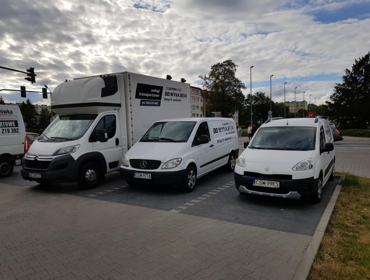 Flota białych samochodów dostawczych z logotypami firmy transportowej ze Świecia, zaparkowana na szarym brukowanym parkingu w pochmurny dzień.