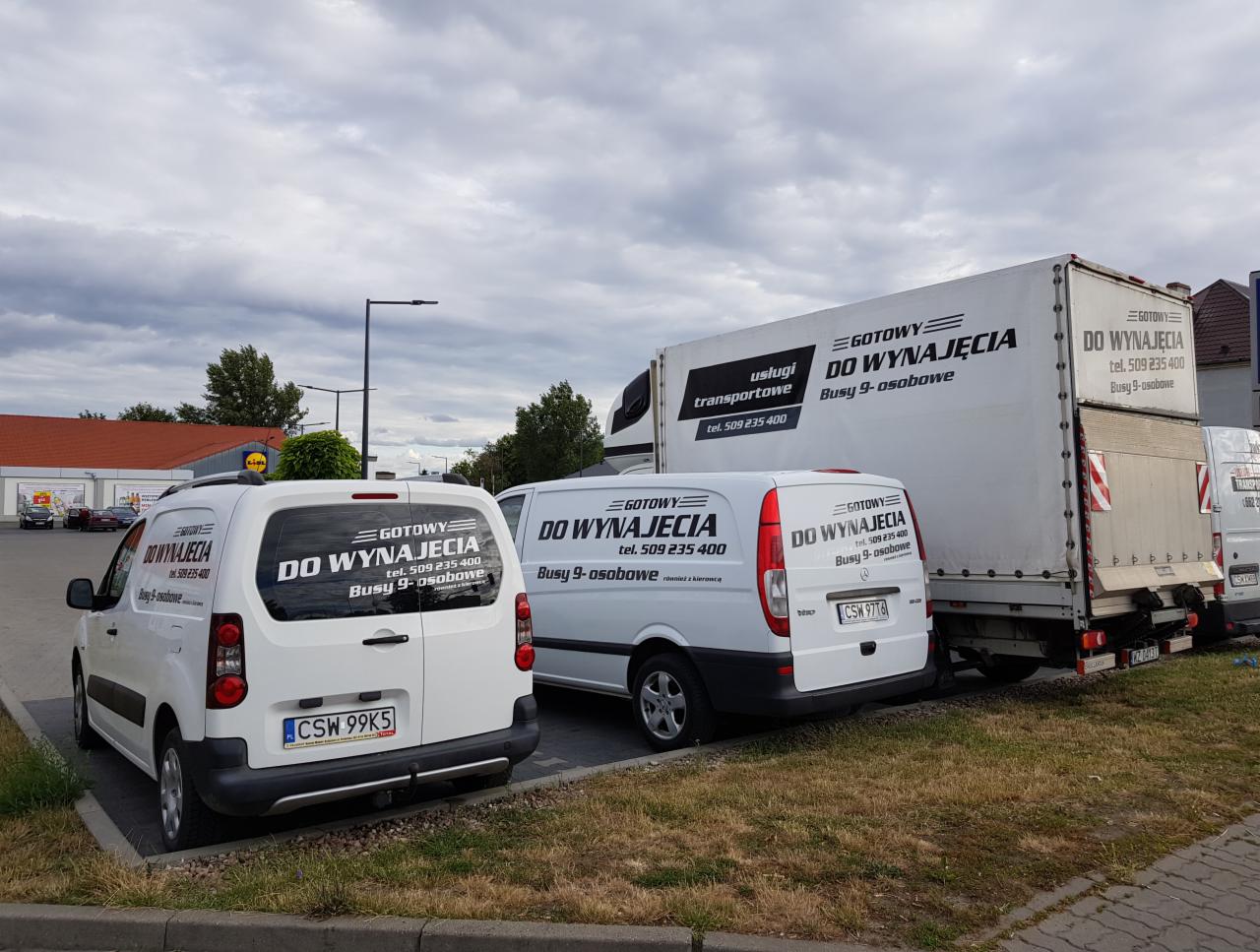 Flota pojazdów firmy transportowej: biały bus, furgonetka i ciężarówka z logo 'GOTOWY DO WYNAJĘCIA, Busy 9-osobowe' zaparkowane na parkingu.